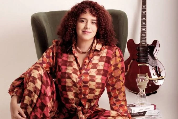 A woman with curly red hair wearing a multicolored geometric patterned outfit sitting in a green armchair with a guitar and stack of books nearby.