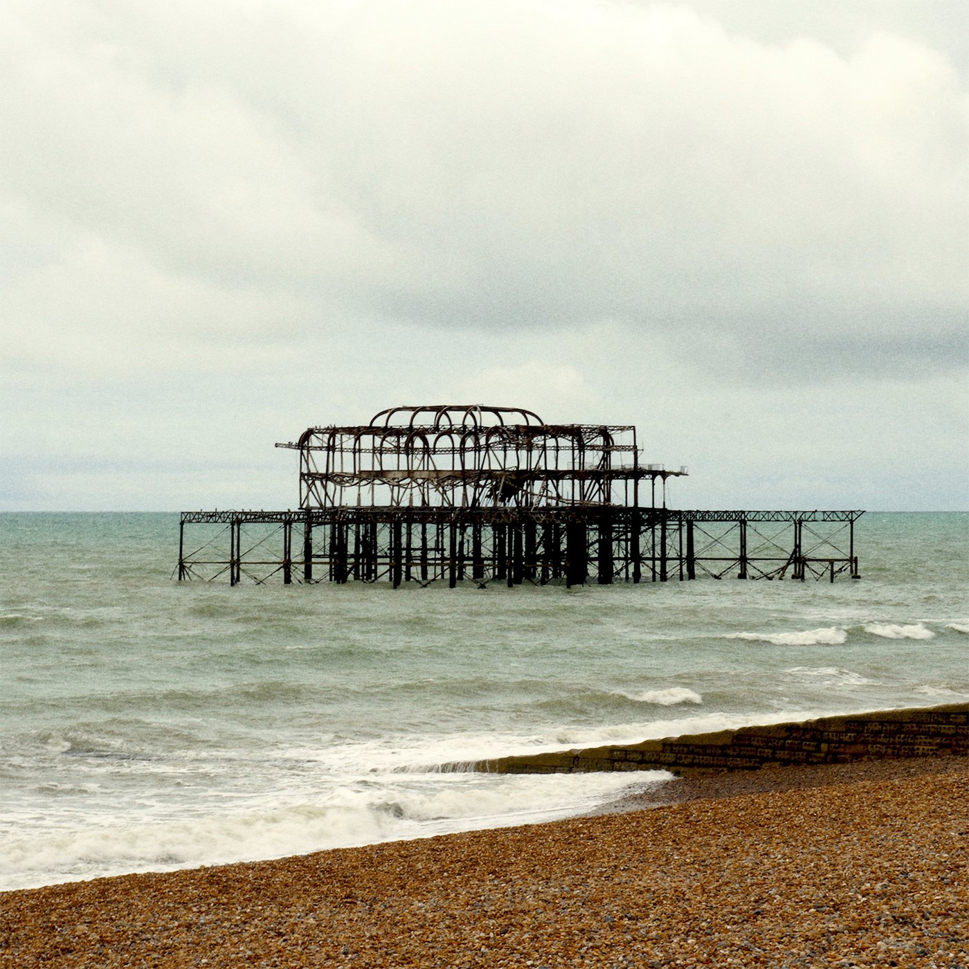 An abandoned, rusted seaside pier with missing sections, extending into the ocean under cloudy skies.