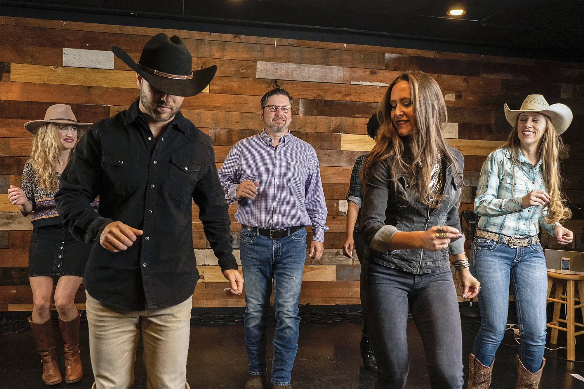 Group of five people line dancing to Cajun music in a rustic venue with wood walls, including two women wearing cowboy hats and cowboy boots, and two men, one with a cowboy hat.