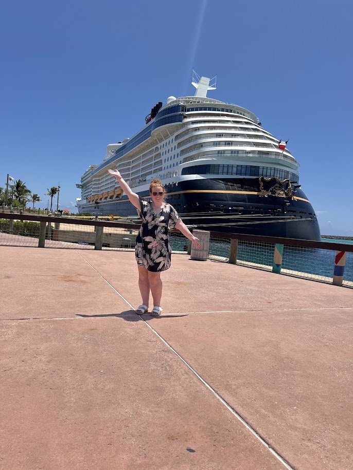 A woman in a floral dress standing on a dock with a cruise ship in the background, waving at the camera on a sunny day.