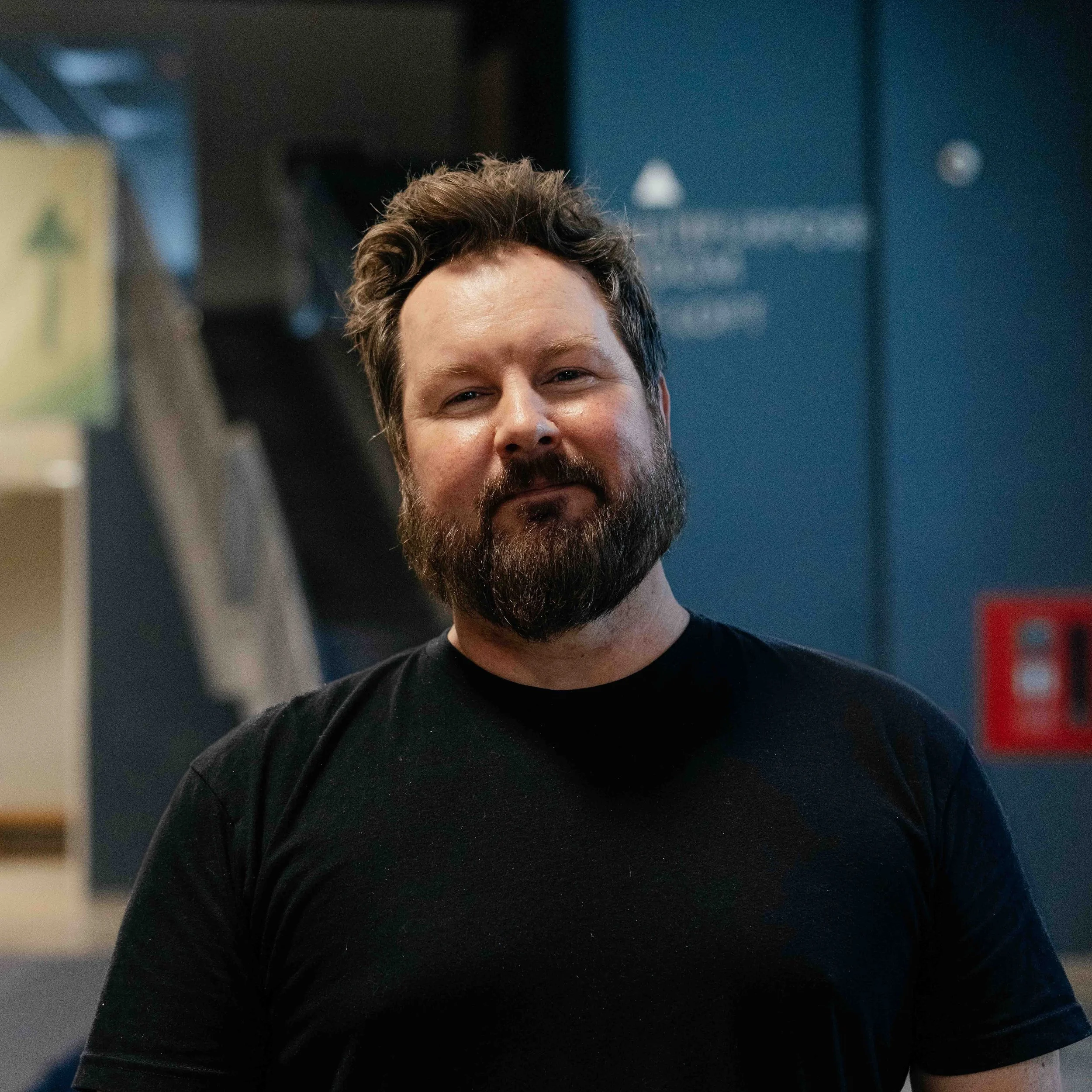 A smiling man with glasses and short dark hair, wearing a gray patterned shirt, standing indoors with a blurred background.
