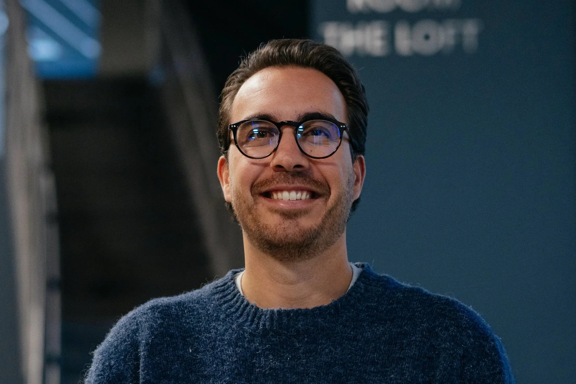A man with glasses and a beard smiling indoors.