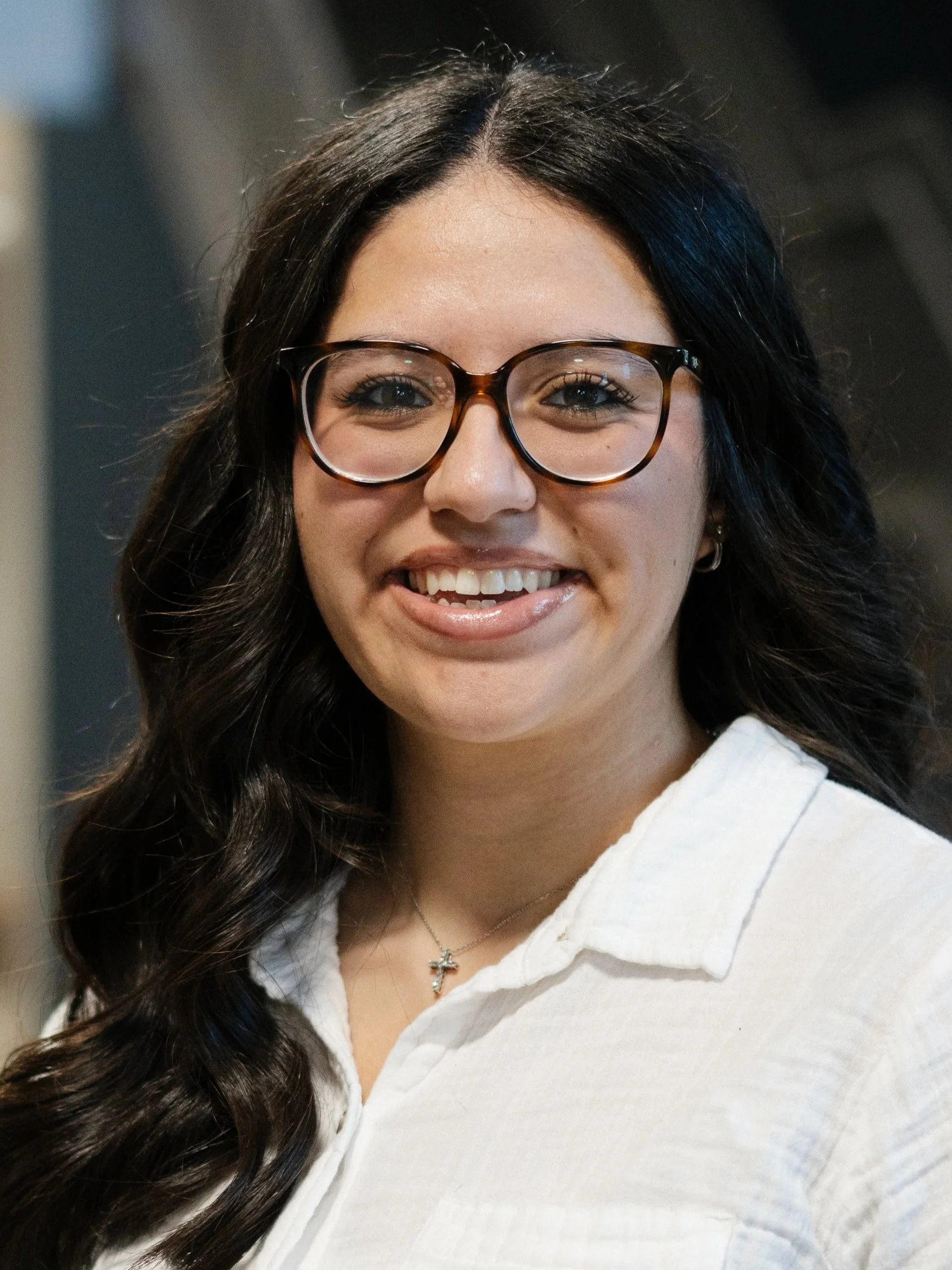 Smiling woman with dark wavy hair, glasses, and earrings, indoors with blurred background.