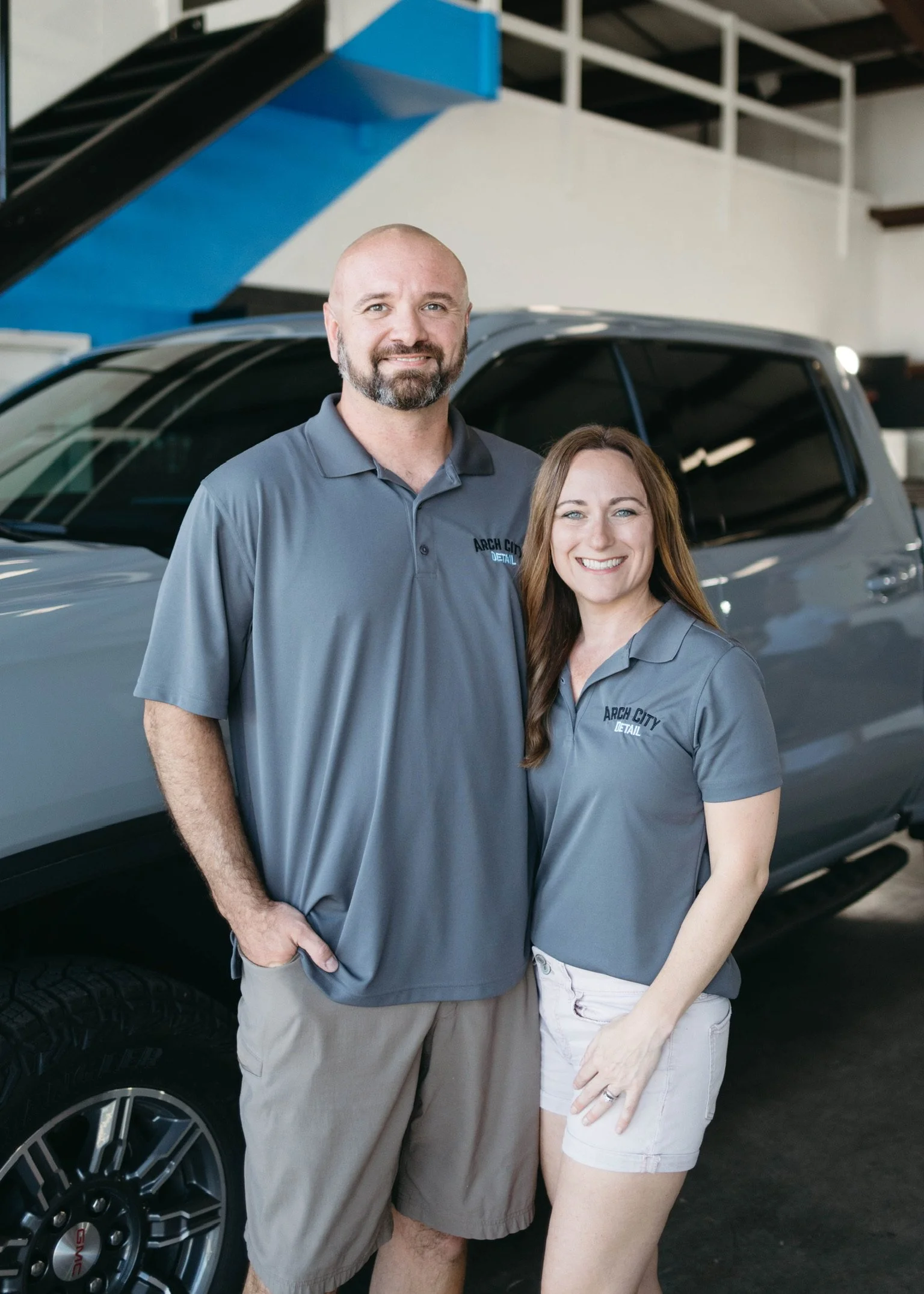 A man and woman standing in front of a gray vehicle inside a garage, both wearing matching gray shirts with 'Arch City Detail' logo, smiling at the camera.