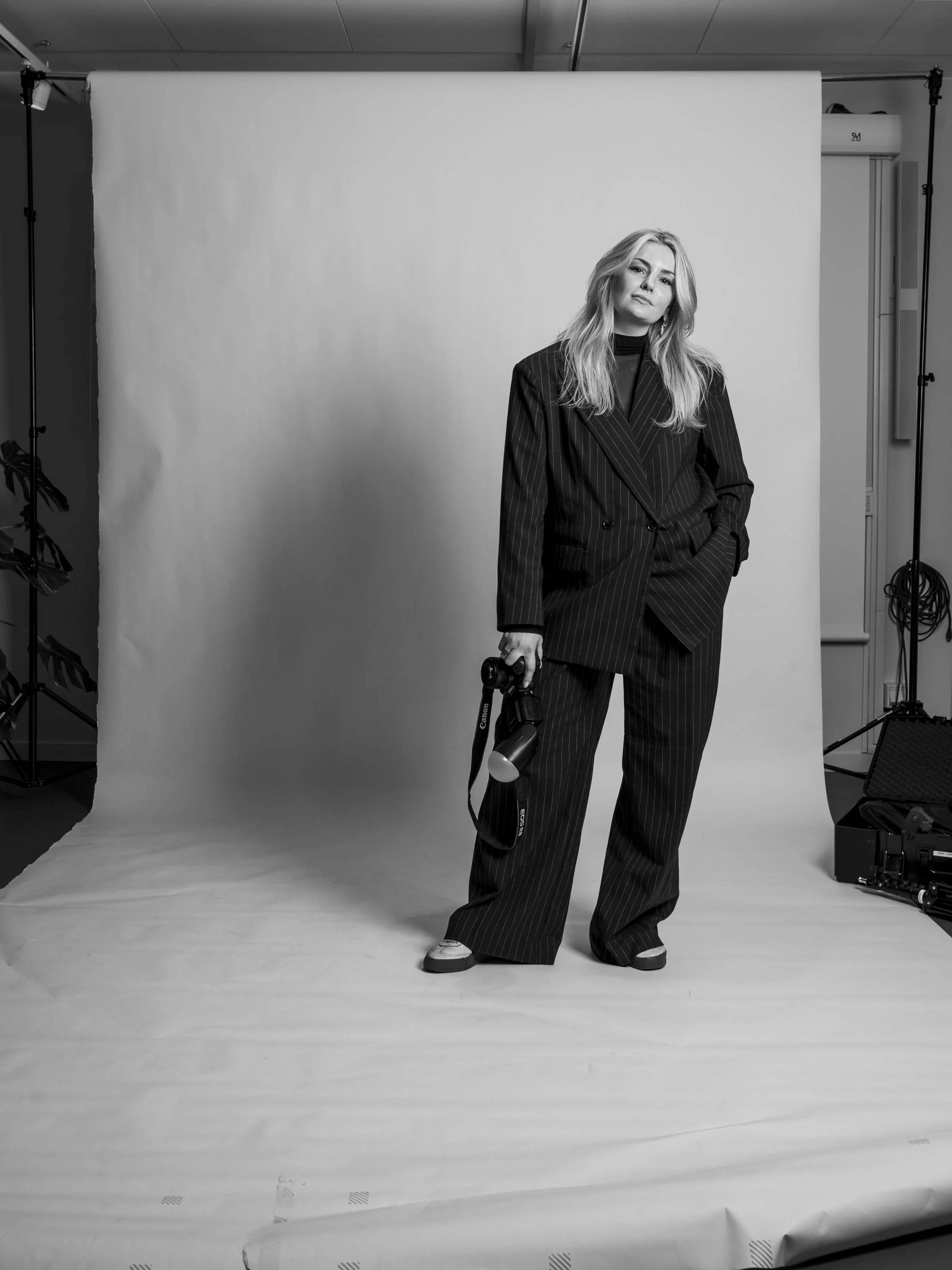 En kvinna står framför en vit bakgrund i ett fotostudio. Hon bär en mörk kostym med randiga linjer och håller en kamera i handen. Fotograf i Jönköping för privat och Företag. Fotografi, Fotograf