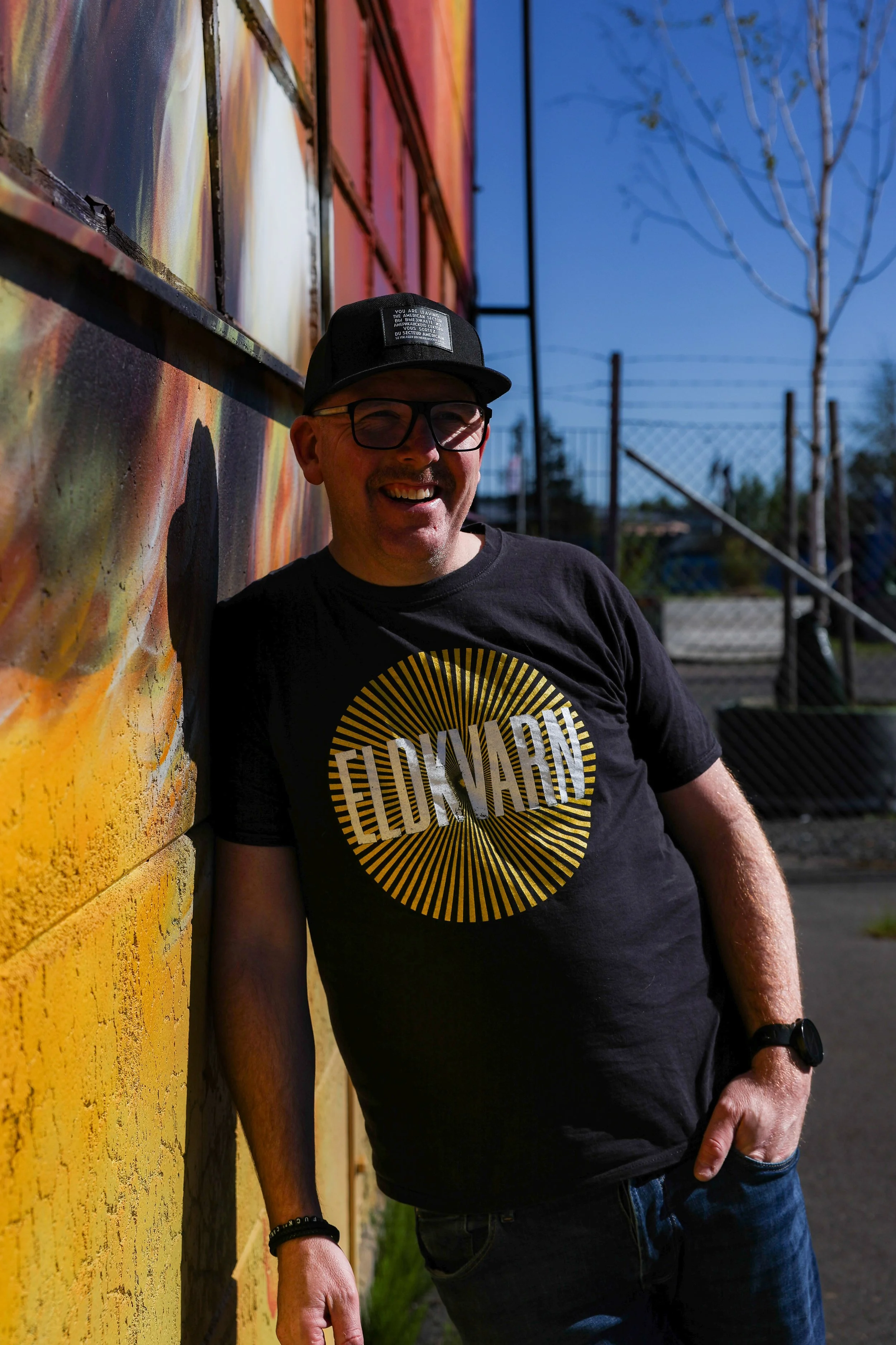 En glad man med glasögon, rak mustasch, svart keps och svart t-shirt poserar bredvid en gul vägg med graffiti, på en solig dag.
