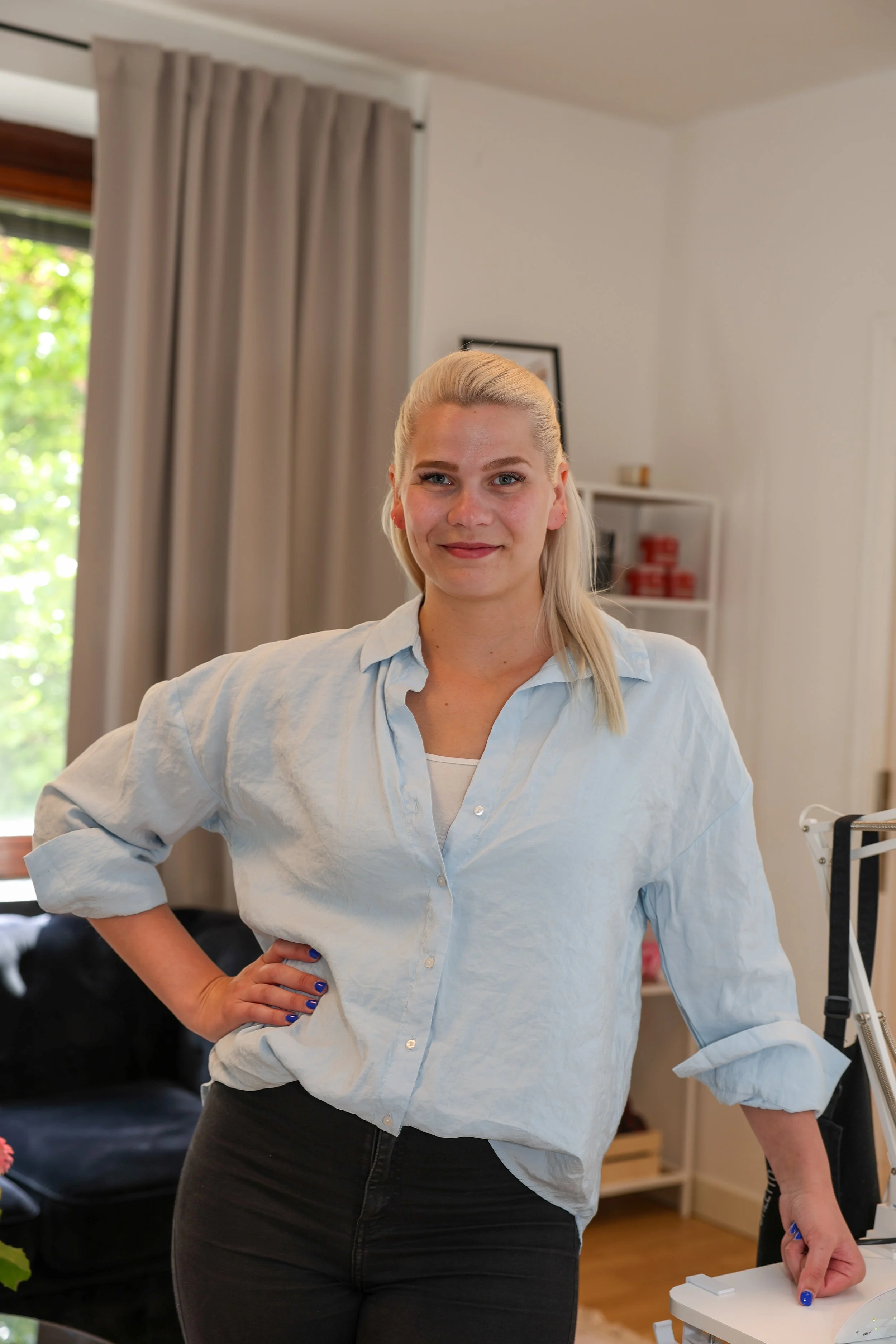 En ung kvinna med blont hår, iklädd vit skjorta och svarta jeans, står i ett inomhusrum. Hon ser direkt mot kameran och ler lätt. Bakom henne finns ett fönster med ljusa gardiner och en vit hylla med några föremål.