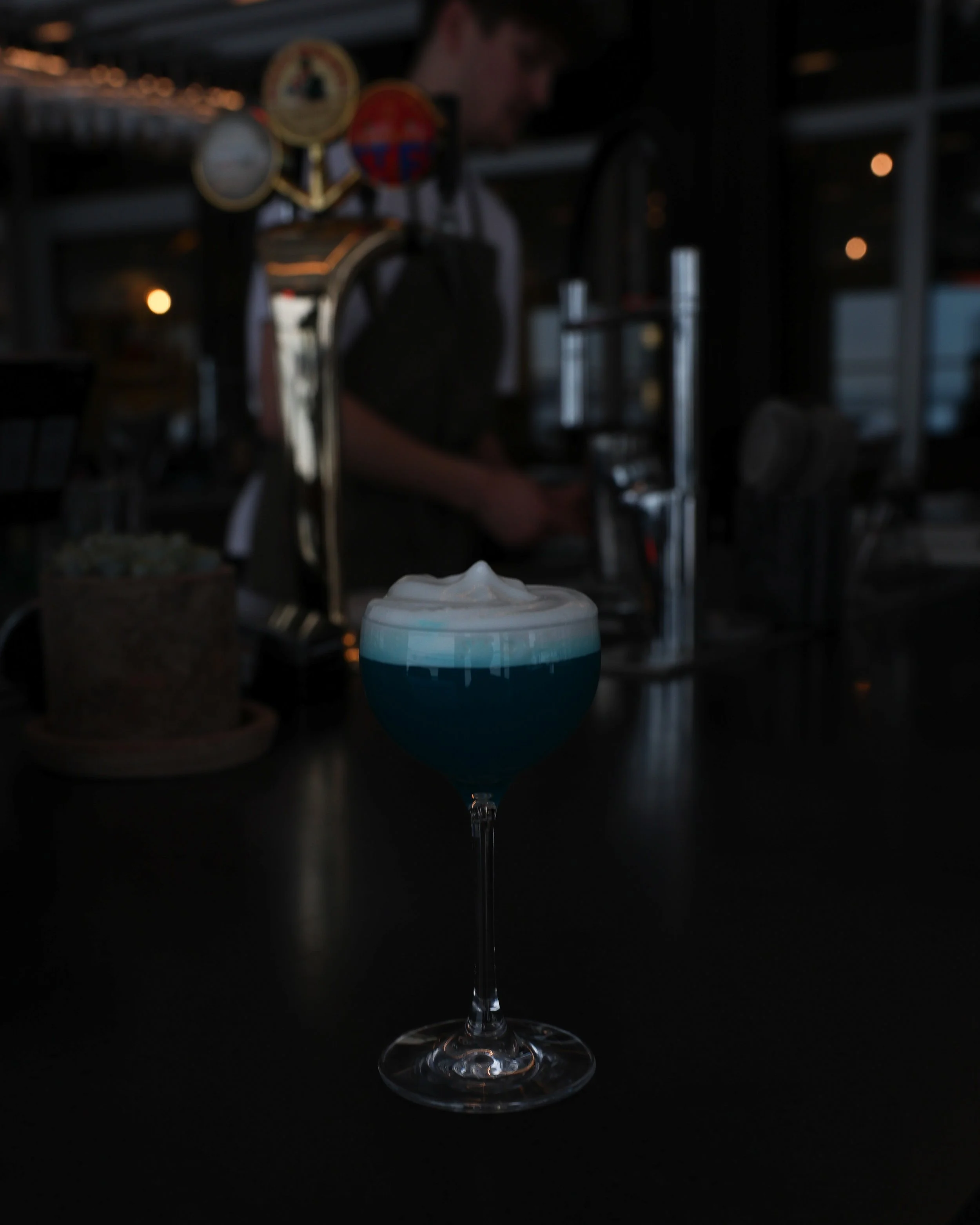 En blå cocktail i ett vinglas med grädde på toppen, placerad på ett svart bord. Bakgrund är mörk med några utblåsta ljuspunkter och skuggfigurer, inklusive en person i vit t-shirt.