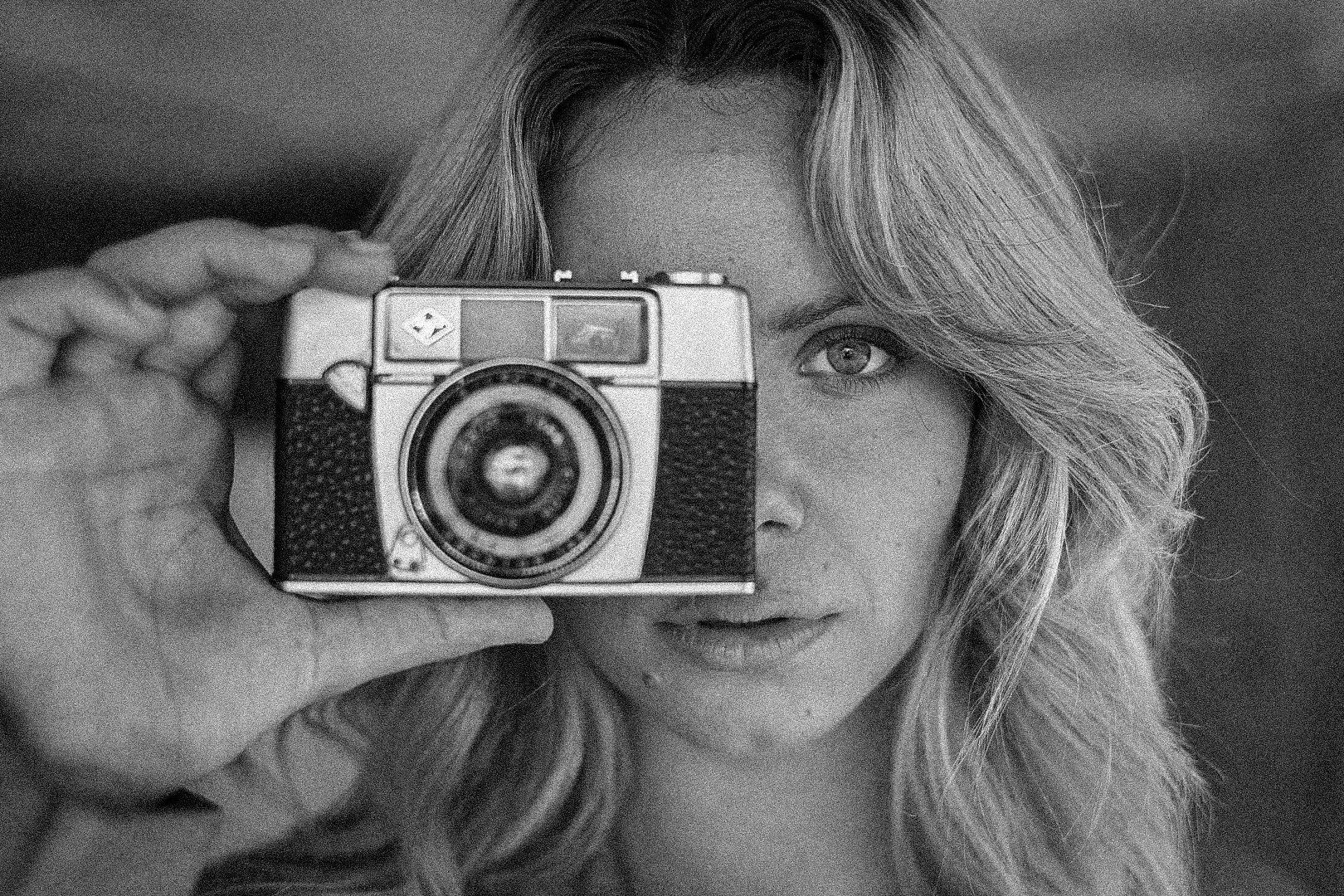 Svartvitt foto av en ung kvinna med blont, lockigt hår som tar ett självporträtt med en vintagekamera.Jönköping modellfotografering av Nathalie Höglin