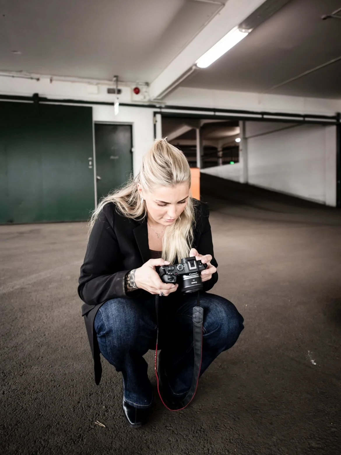 Kvinna med blont hår i svart jacka och jeans knoppar ner för att titta på en kamera i ett parkeringsgarage. Jönköping företagsfoto av Nathalie Höglin