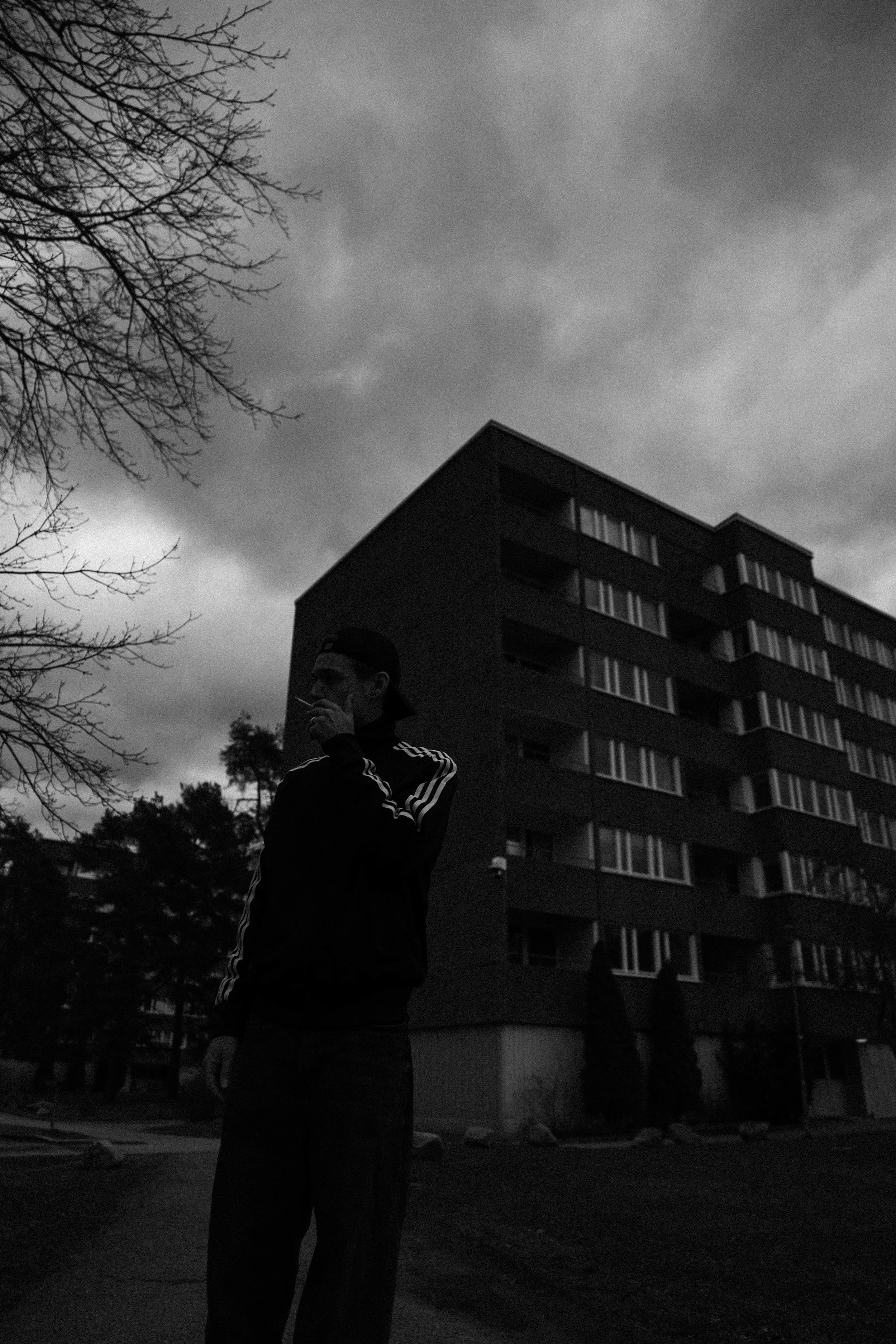 En mörk och molnig stadsskyltning med ett flerbostadshus i bakgrunden. En person i mörka kläder och mössa som röker står i förgrunden.