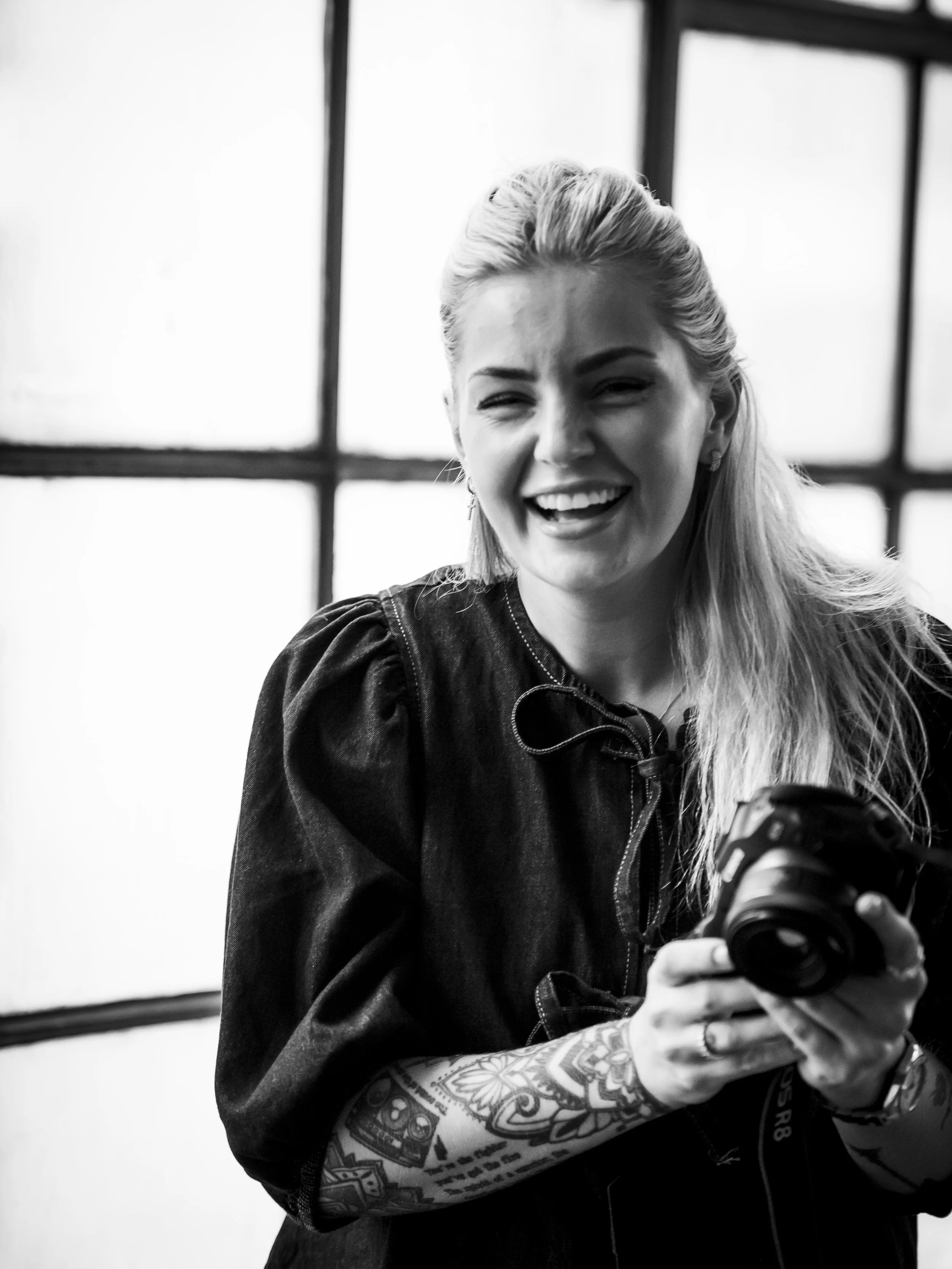 En leende kvinna med tatueringar på armen håller en kamera, i svartvitt. Jönköping företagsfoto av Nathalie Höglin