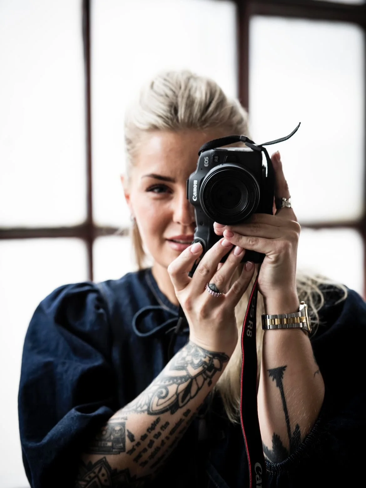 En kvinna med blont hår som fotograferar med en kamera framför ansiktet, mot en bakgrund av stora fönster. Jönköping företagsfoto av Nathalie Höglin