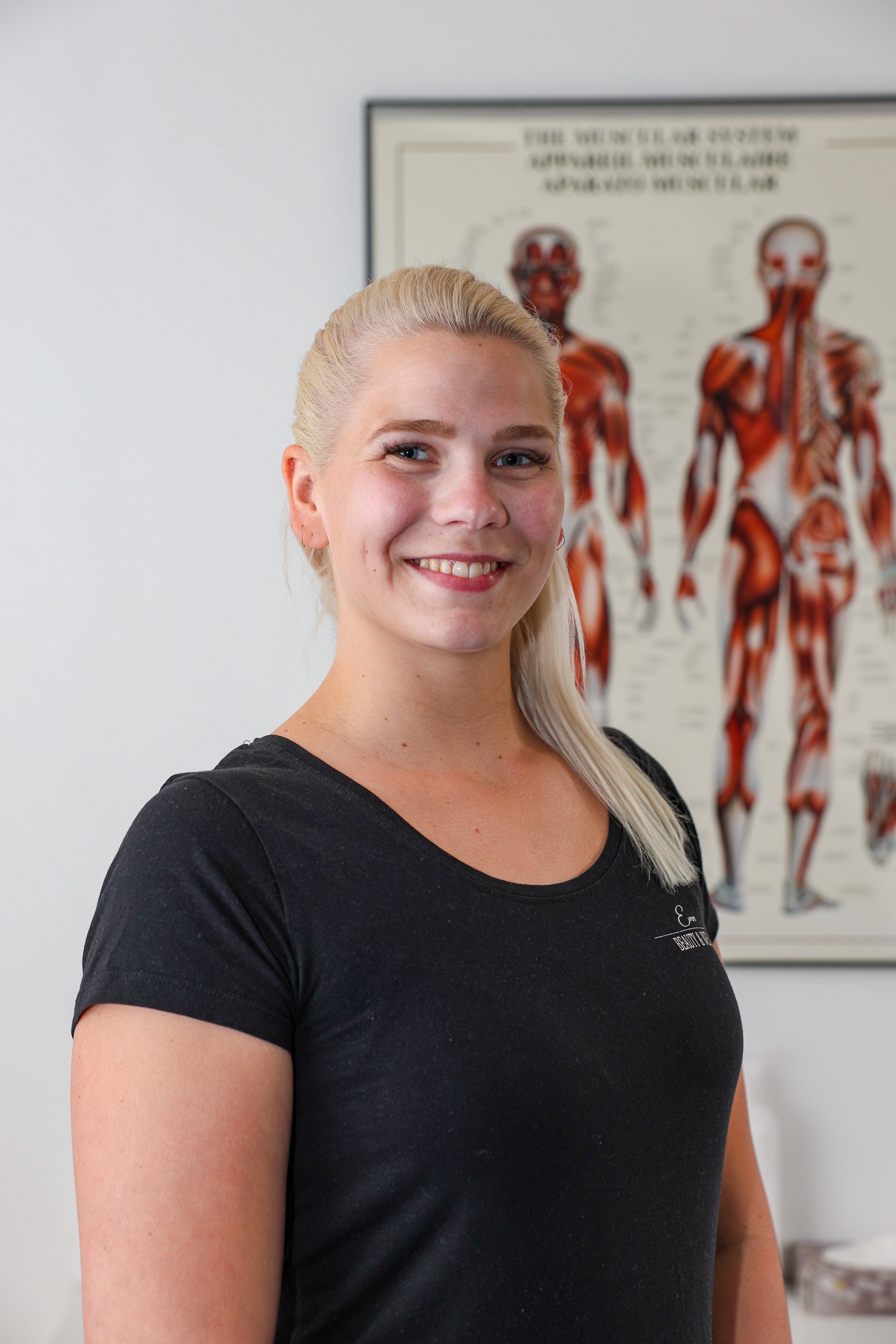 En kvinna med blont hår, leende, svart t-shirt står i ett rum. I bakgrunden syns en human anatomisk affisch.
