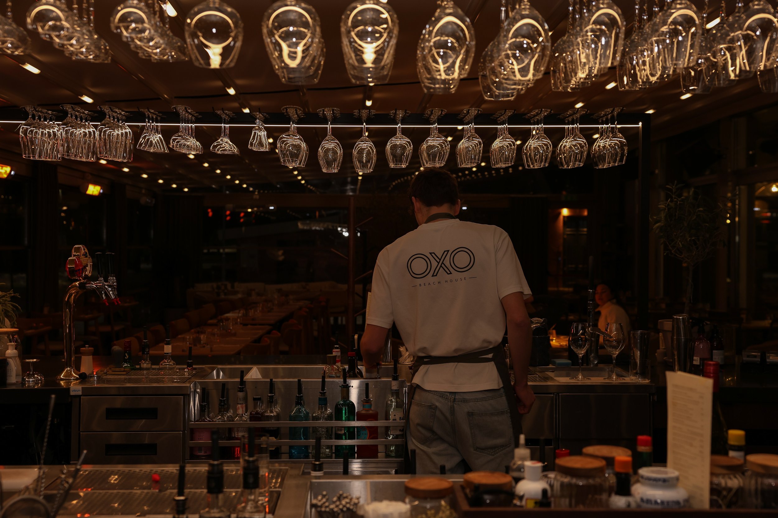 En bartender serverar drinkar bakom en bar i en restaurang med många vinglas hängande från taket.
