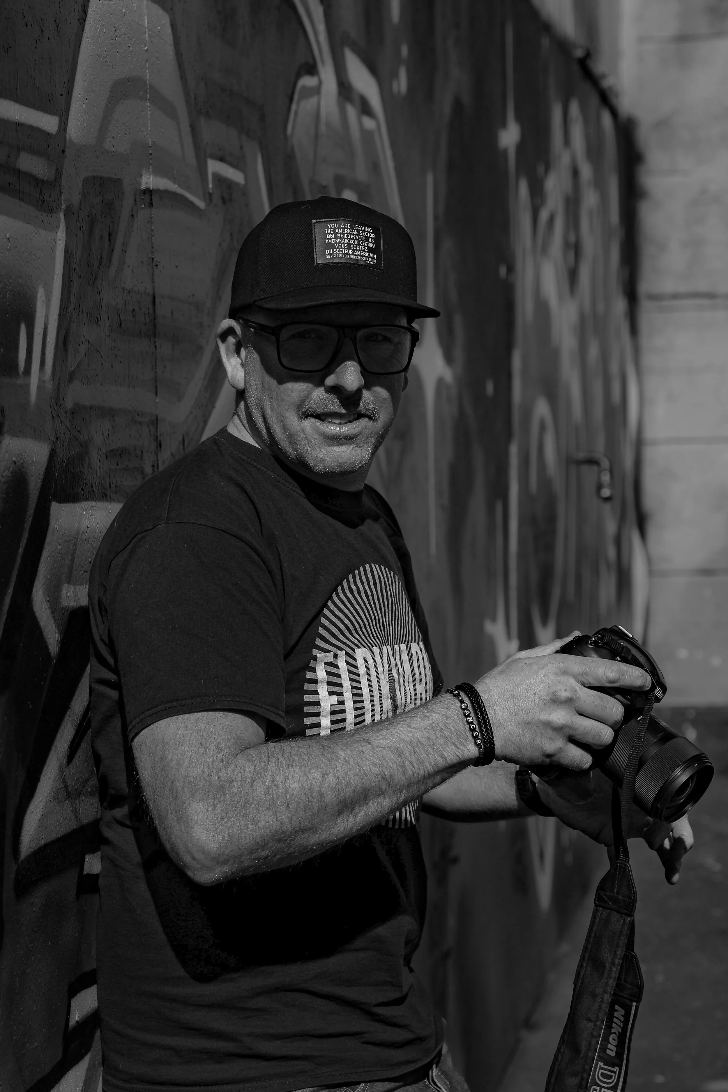 Man med glasögon och keps som håller i en Nikon kamera, står framför en graffiti-adderad vägg.