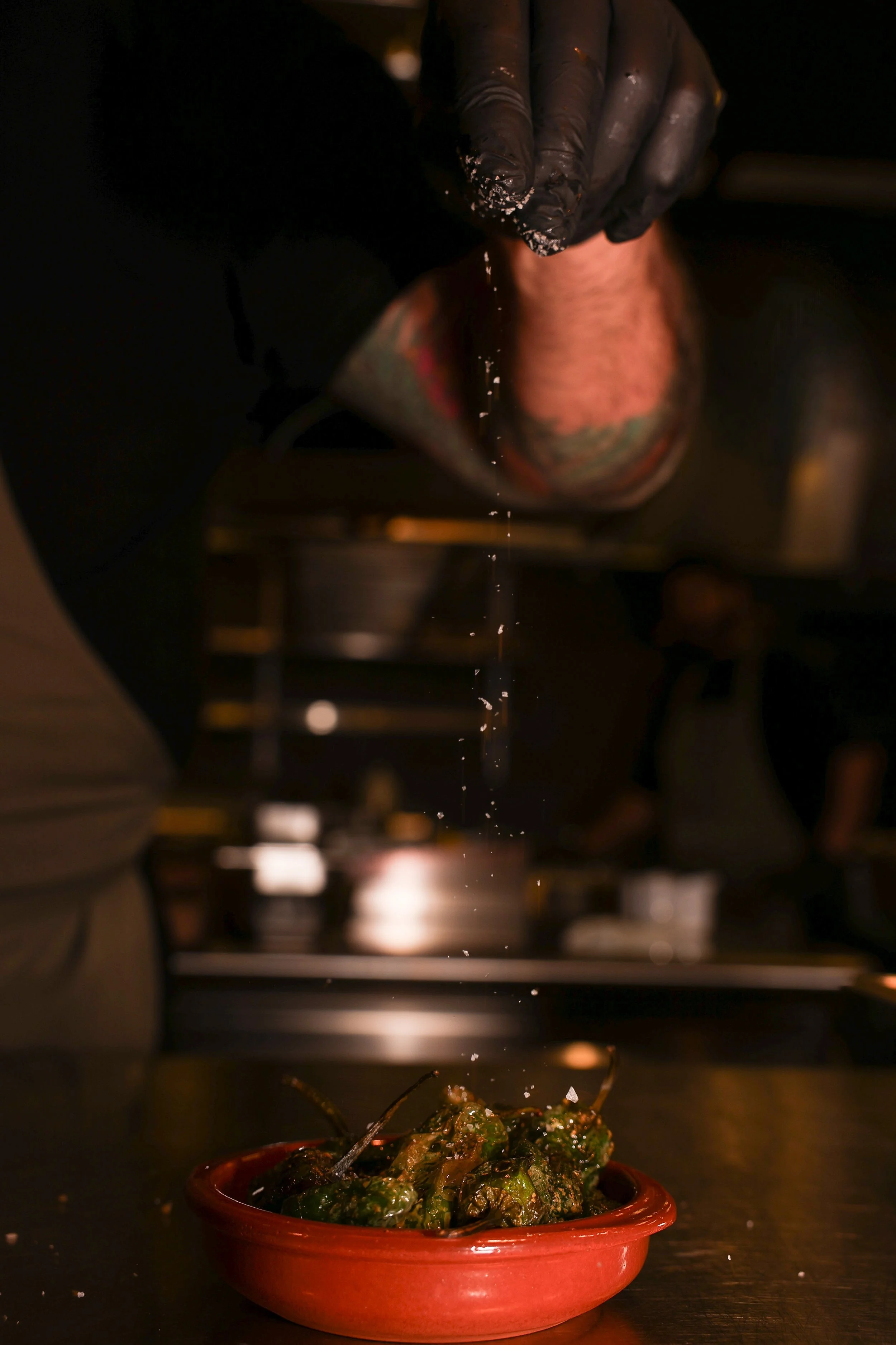 En hand med svart handskar strör salt på en tallrik med grillade grönsaker, under ett ljus.