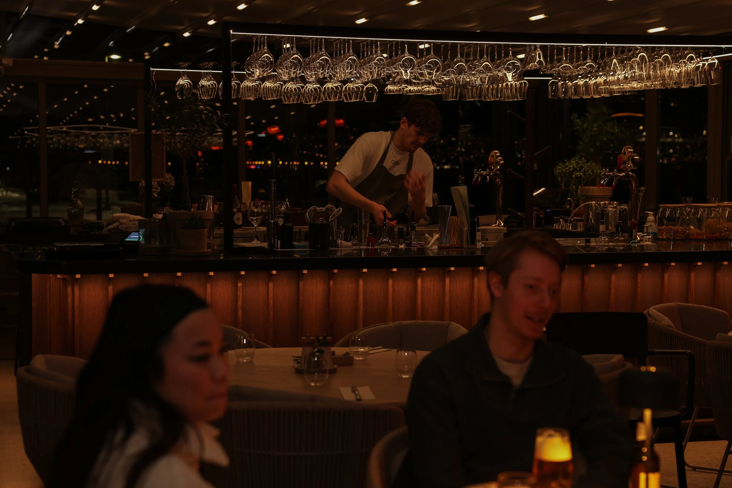 En bartender förbereder drinkar bakom en bar i en restaurang eller bar, med glas hängt uppochner ovanför baren, det är kvällstid och flera personer äter och dricker i restaurangen.