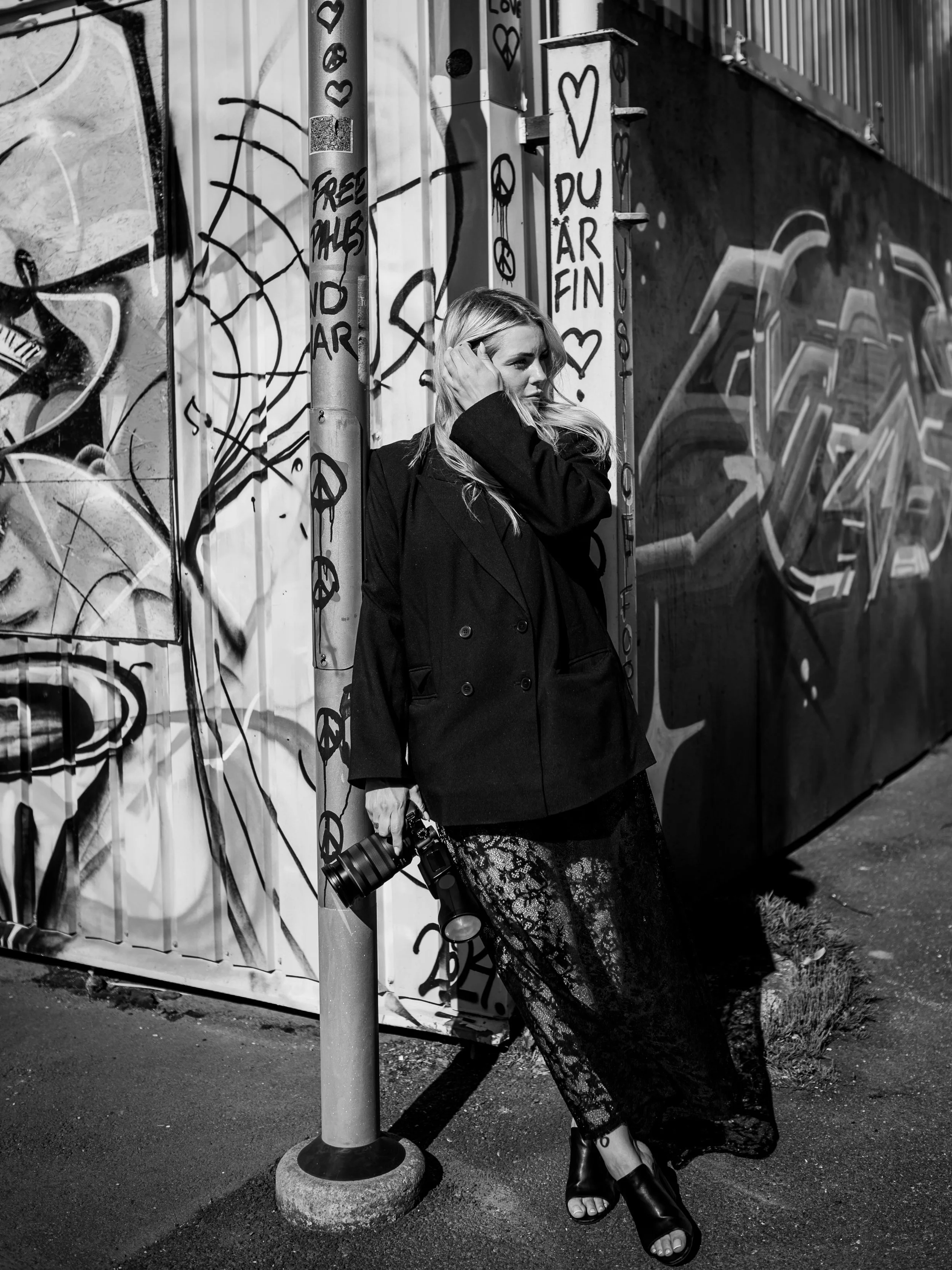 En ung kvinna står mot en graffitiverkad vägg, håller en kameraväska och ser till vänster. Hon är klädd i en svart kappa, lacebyxor och högklackade stövlar, och har långt blont, lockigt hår.