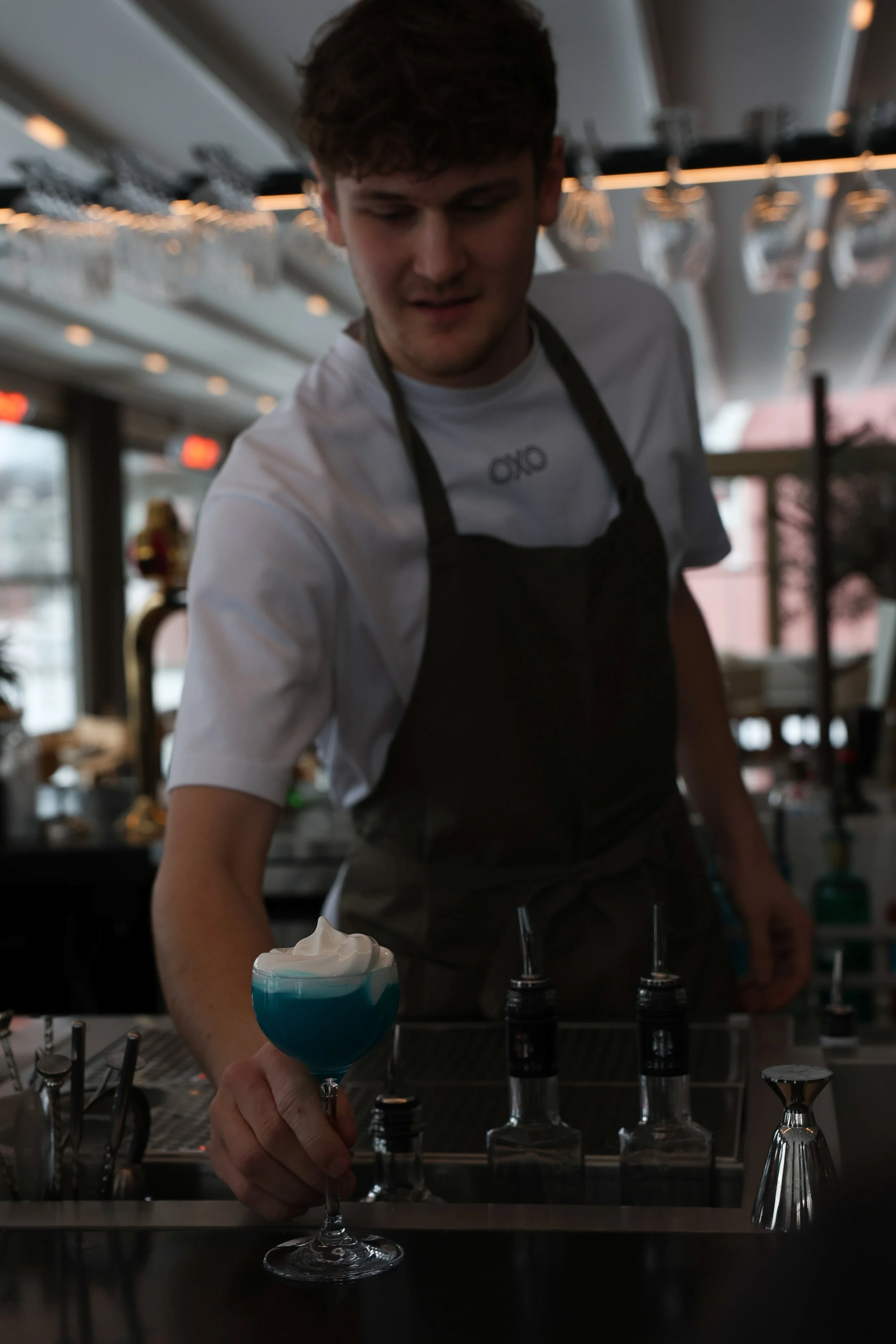 En ung man på en barservering rör vid ett glas med blå och vit dryck, med shaker och flaskor framför sig, och är klädd i en vit t-shirt och svart förkläde, i en modern bar eller restaurangmiljö.