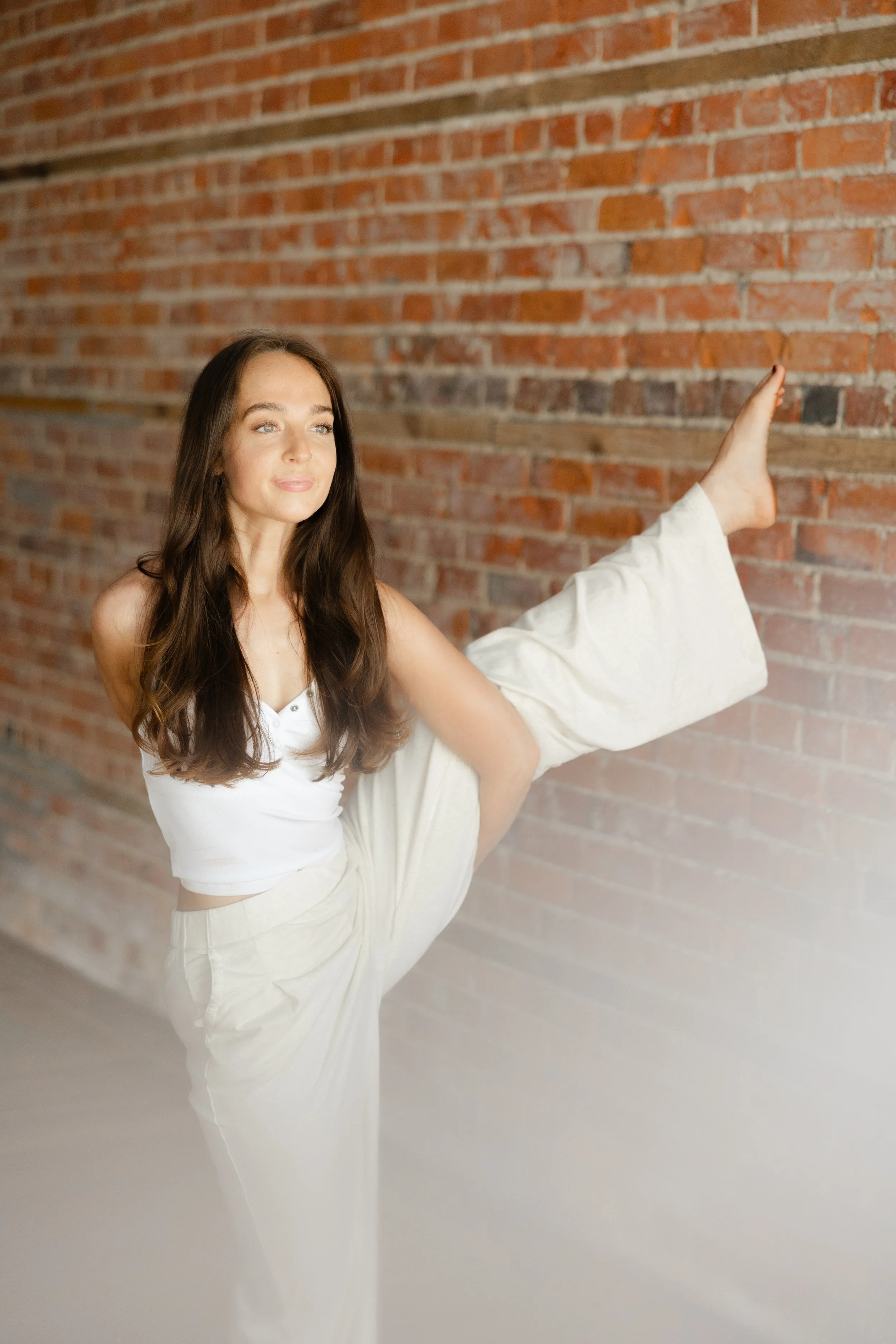 Liz Keeter in a white outfit, in a standing yoga pose in front of a brick wall.
