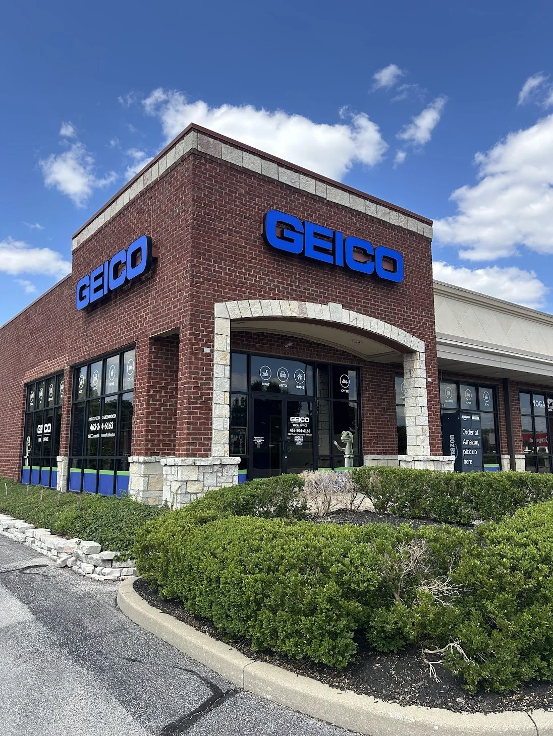 A brick retail storefront with blue 'GEICO' signs and large windows. There are some signs on the windows advertising various insurance services. The building has stone accents around the entrance, and a small landscaped area with bushes in front. The sky is partly cloudy.