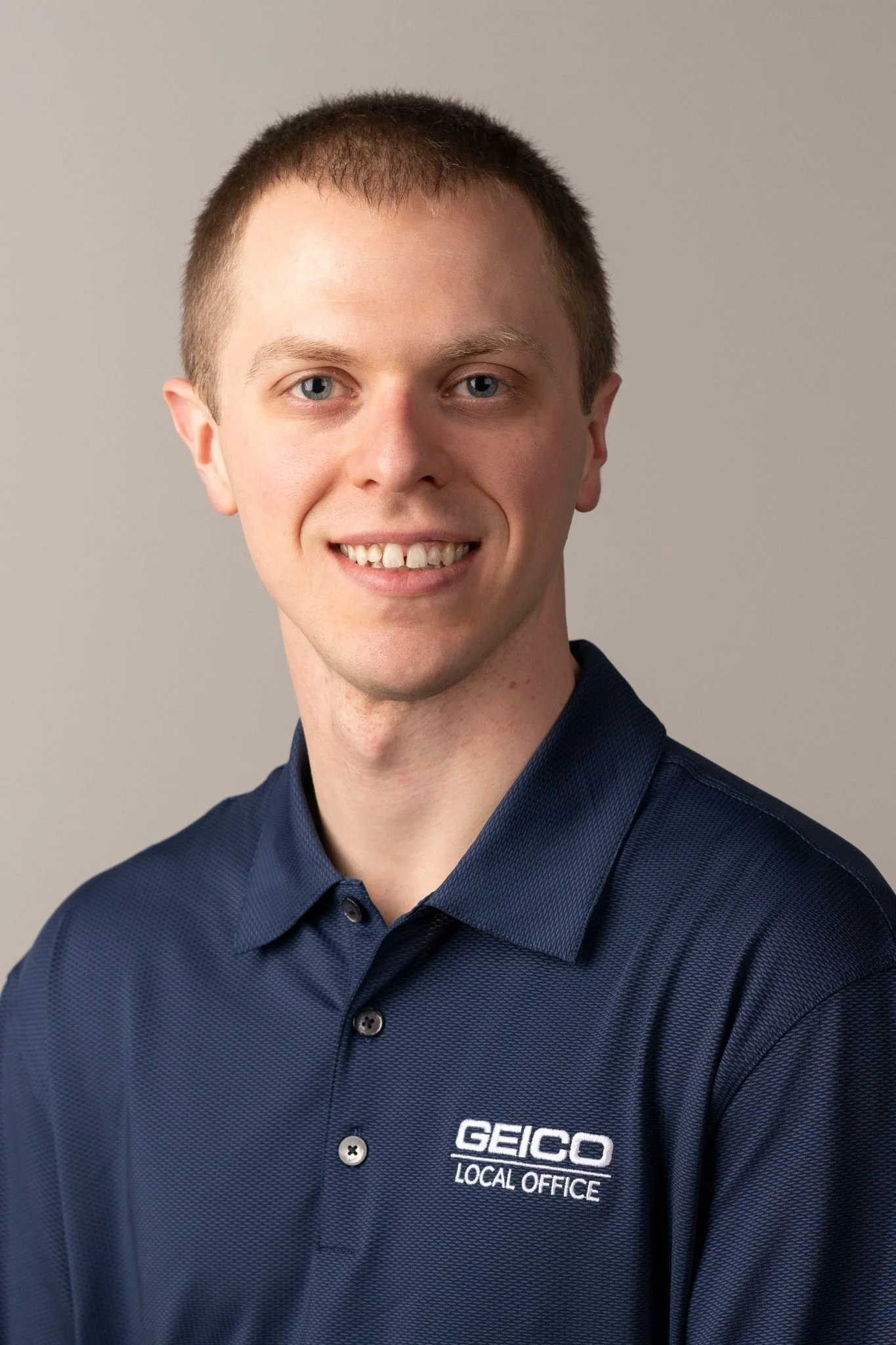 A young man with short brown hair, blue eyes, and light skin, smiling slightly, wearing a navy blue collared shirt with the GEICO logo and the text 'LOCAL OFFICE' embroidered on it, against a plain light gray background.