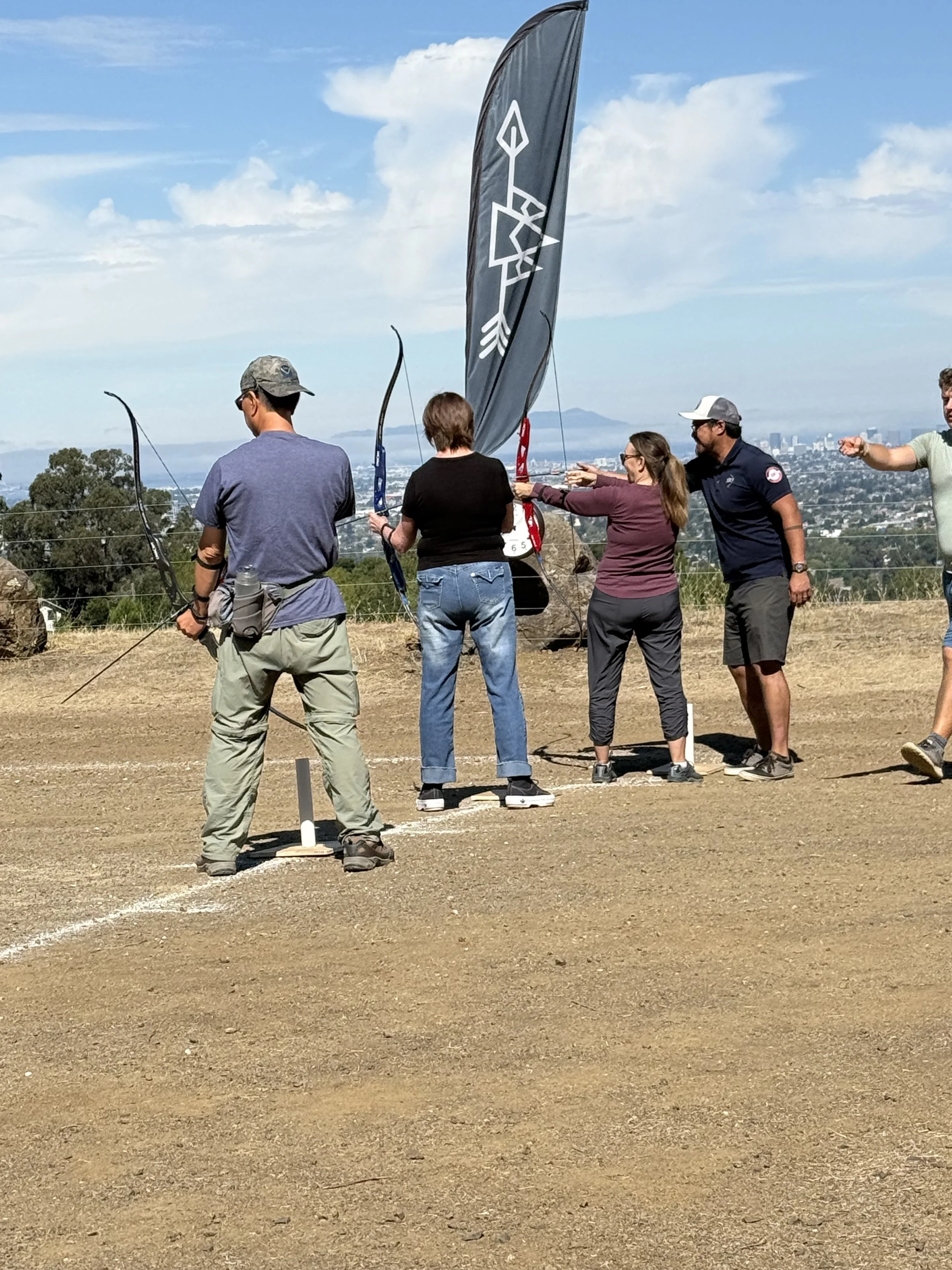 Group of people participating in archery at outdoor field with city skyline in background and a black flag with a white design.