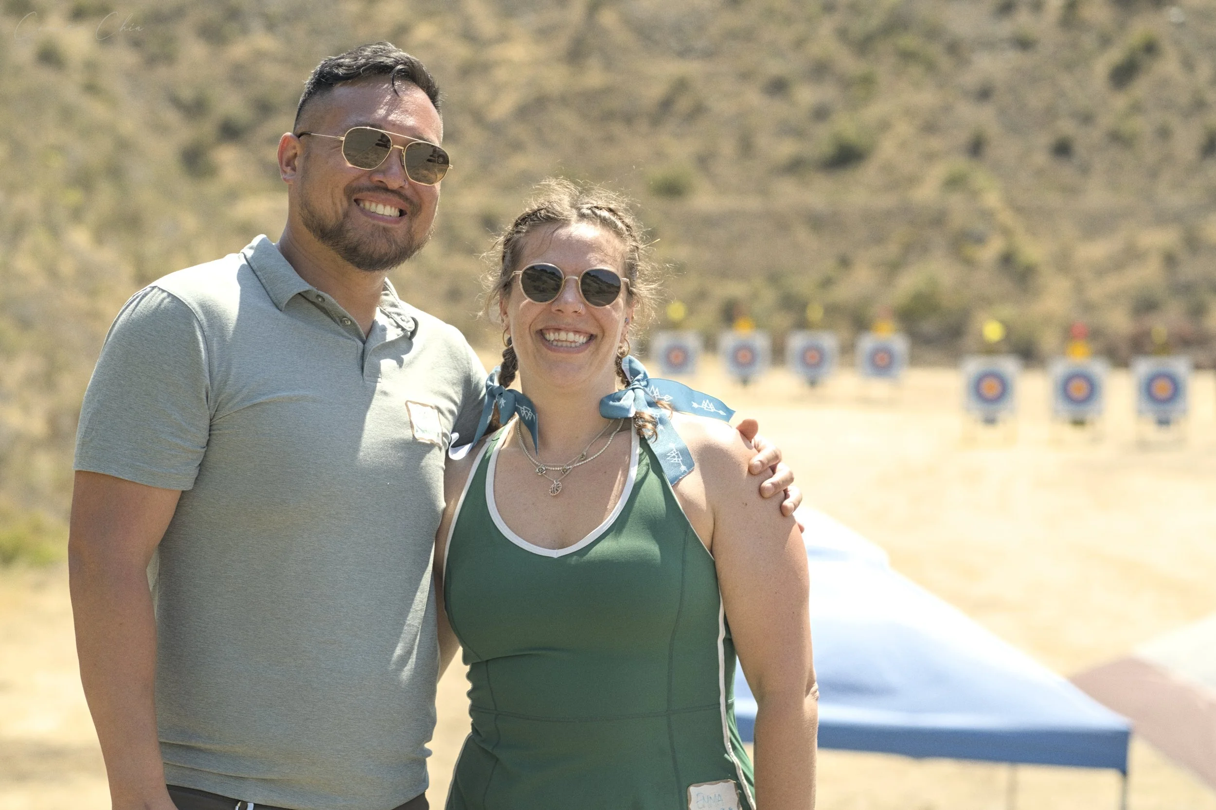 A man and woman smiling outdoors at a shooting range with targets in the background. They are wearing sunglasses and casual summer clothing.