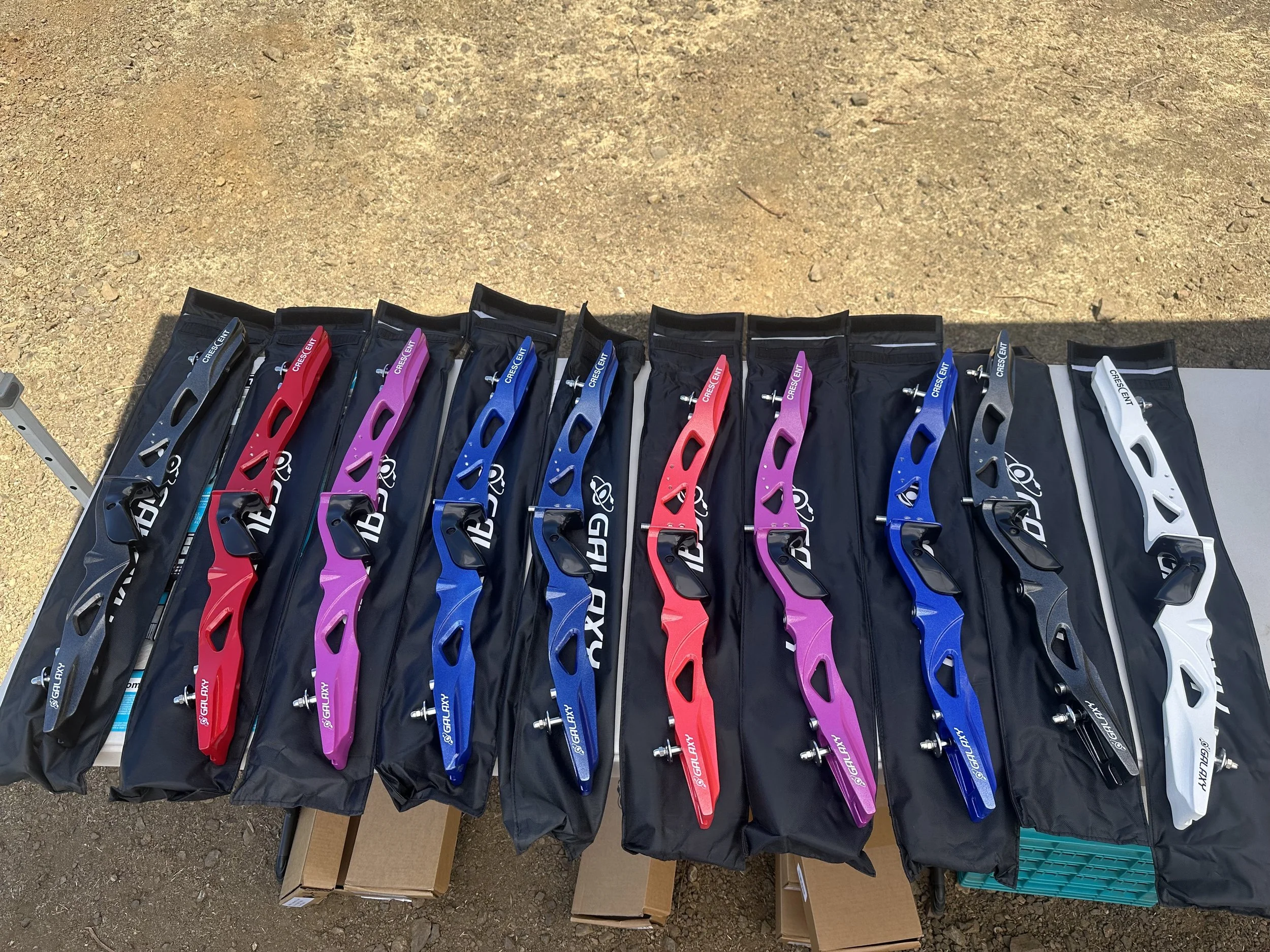 Multiple colorful archery bows laid out on a black tablecloth on a table, with packaging and boxes underneath, outdoors on a dirt surface.