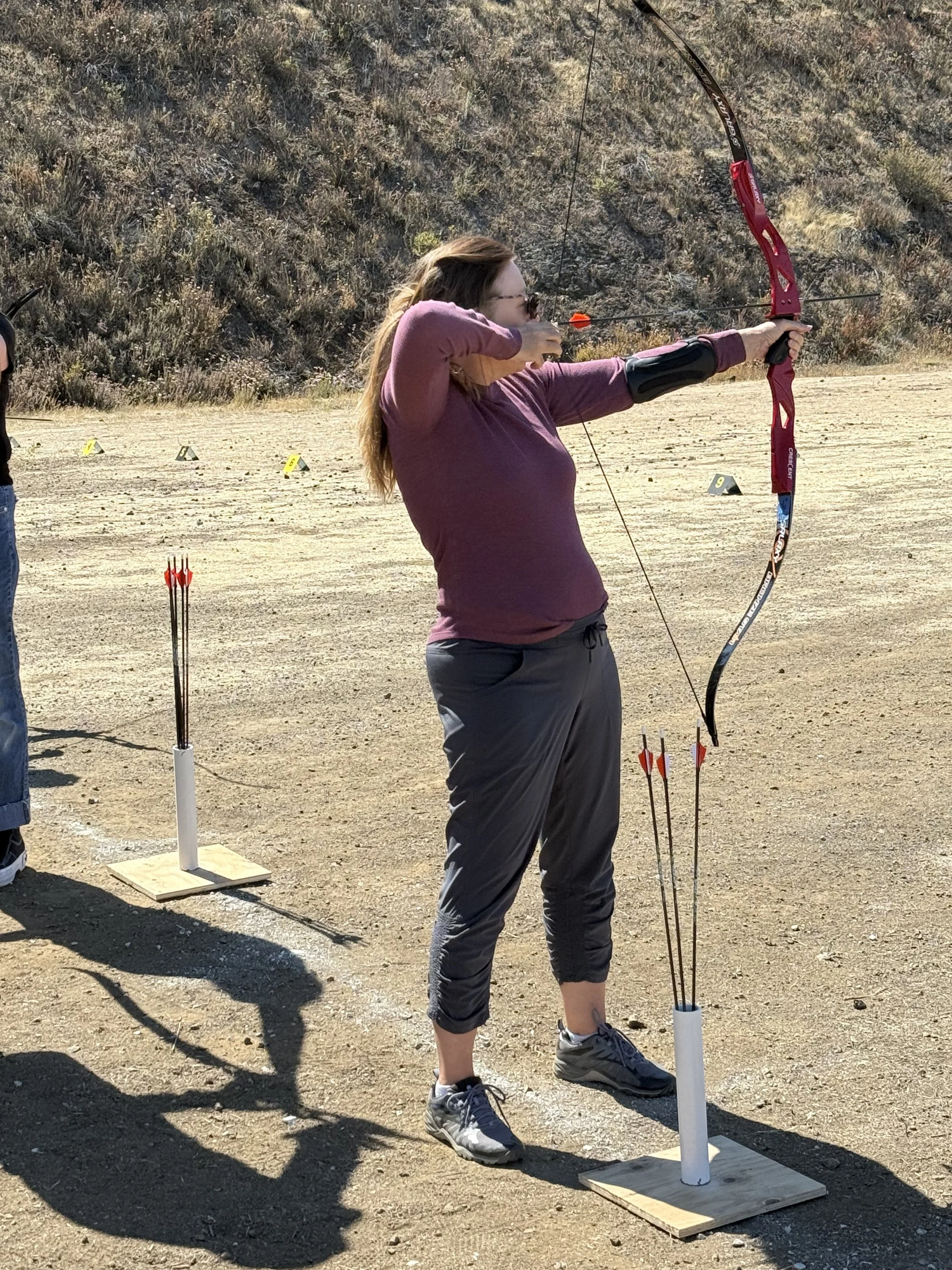 A woman practicing archery outdoors, aiming a bow and arrow at a target in a desert-like area with markers on the ground.
