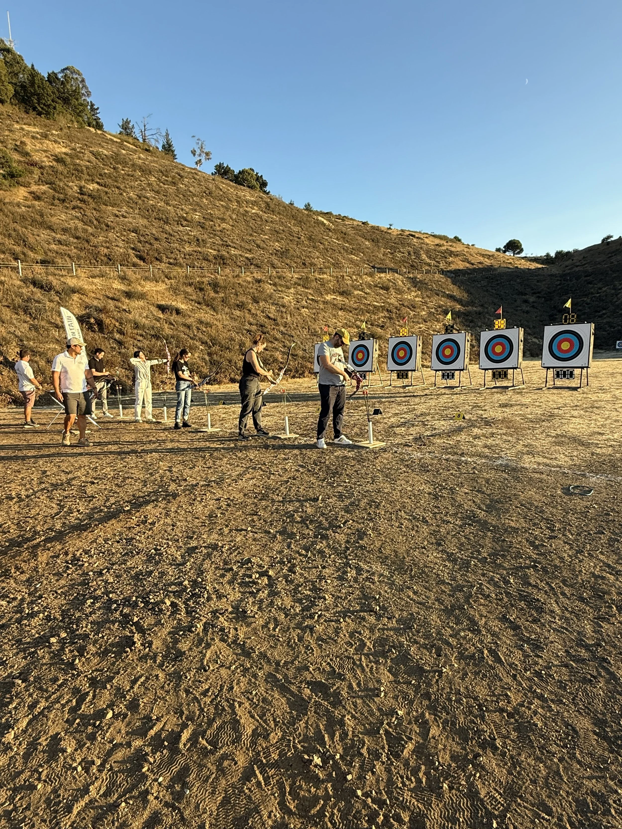 Group of people participating in an outdoor archery activity at a target range on a sunny day, with hills in the background.
