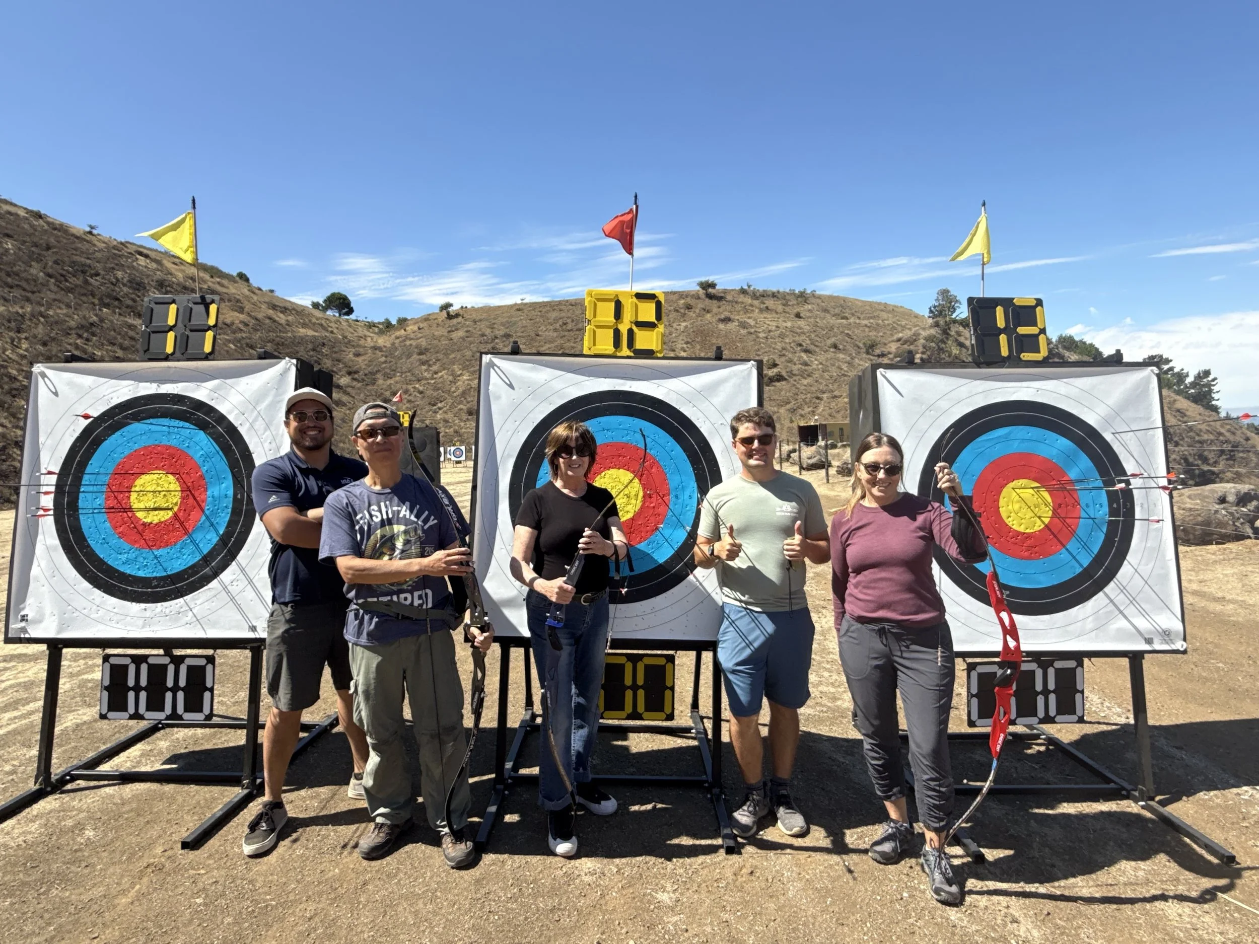 Five people standing in front of three archery targets with bows and arrows, holding equipment and smiling at an outdoor archery range on a sunny day.