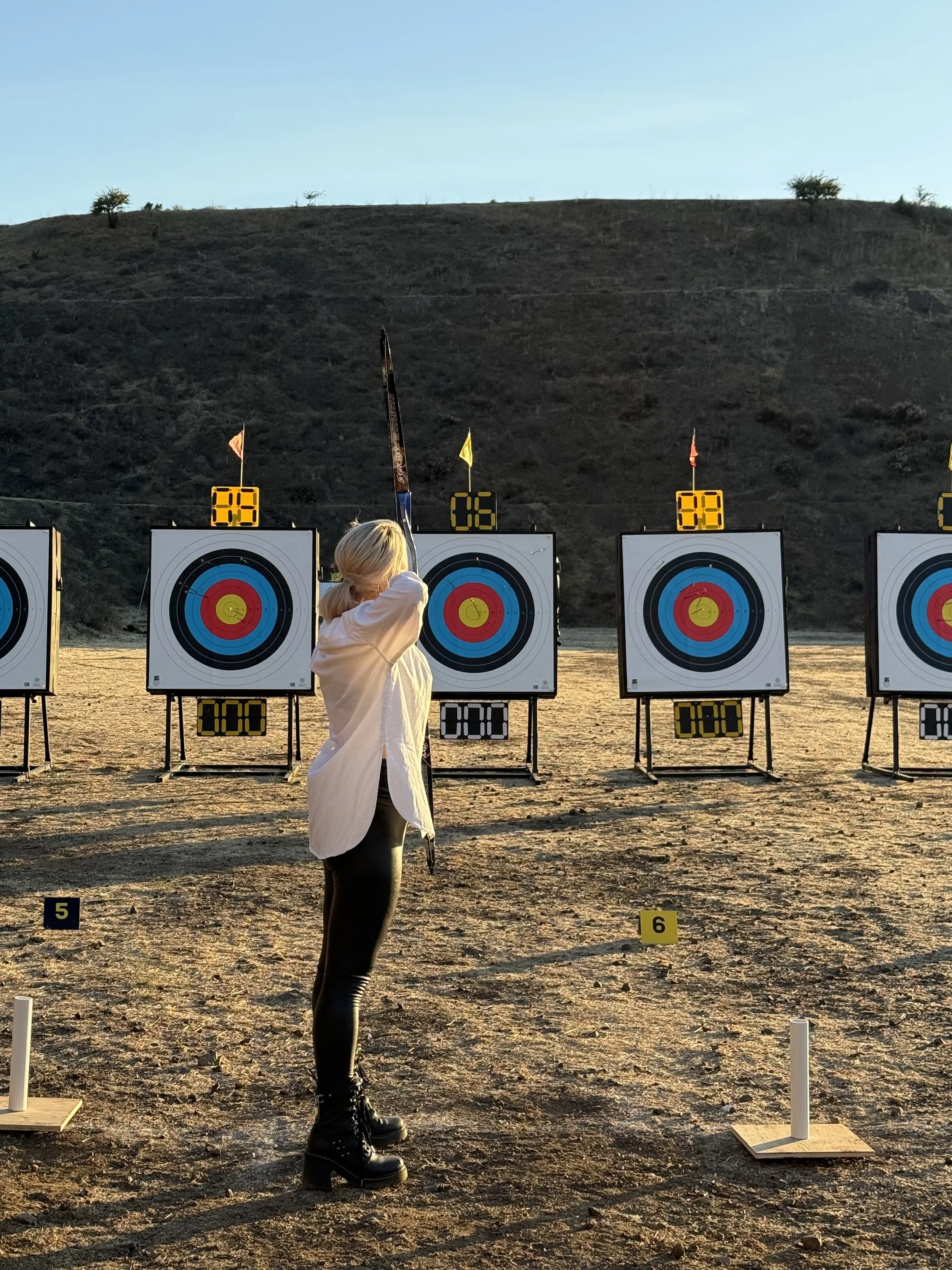 A woman with blonde hair in a ponytail aiming a bow and arrow at an archery target during sunset on a sandy outdoor range.