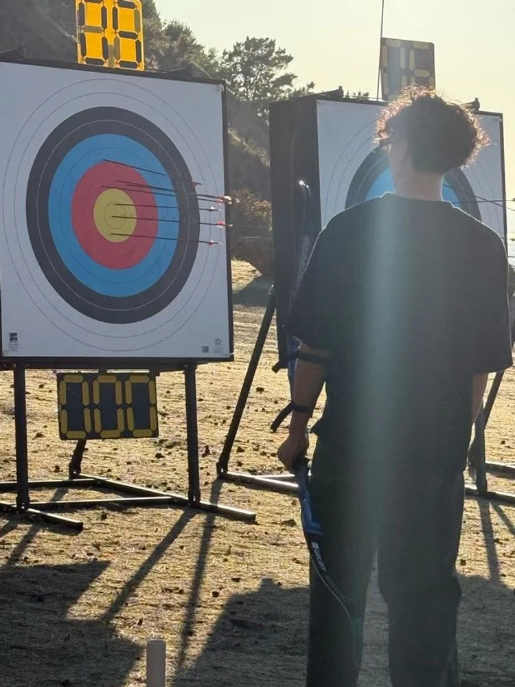 A person with curly hair aiming a bow and arrow at an archery target outdoors during the daytime. The target has multiple colored concentric circles with arrows sticking in the red and yellow sections. A digital timer is visible above the targets.