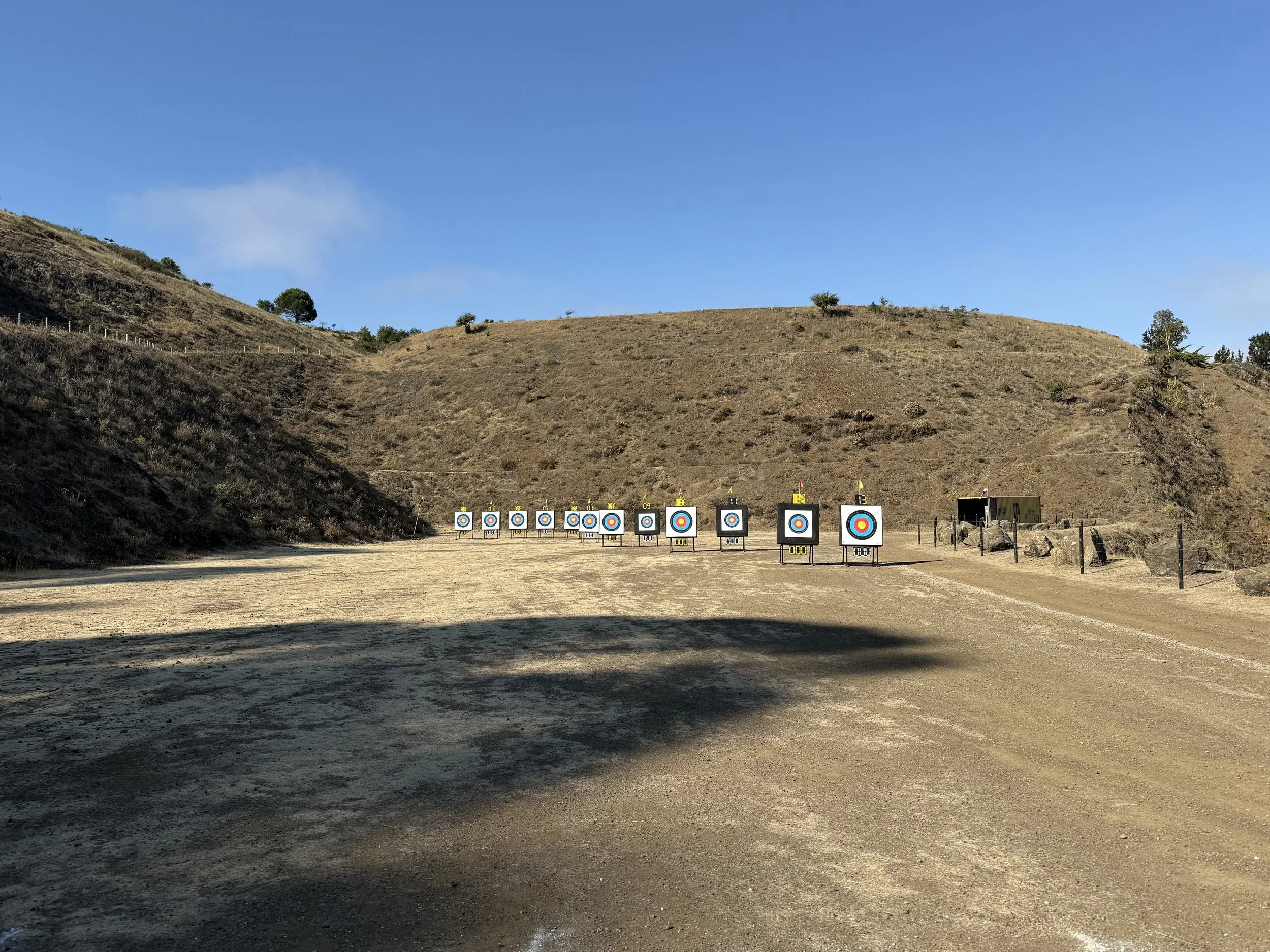 Multiple archery targets set up outdoors on a dirt field, facing a hillside under a blue sky with some clouds.