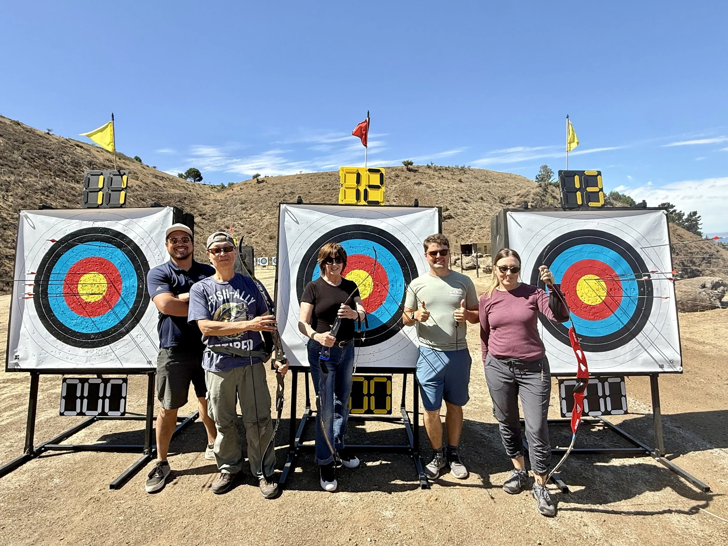 Group of five people standing in front of archery targets outdoors on a sunny day, with hills in the background. They are holding bows and arrows, and some are giving thumbs up.