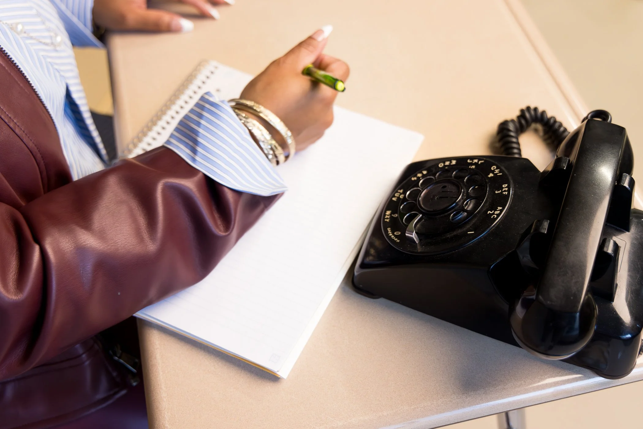 A person wearing a leather jacket and blue striped shirt is writing on a notepad with a green pen. An old-fashioned black rotary telephone is placed on the desk next to them.