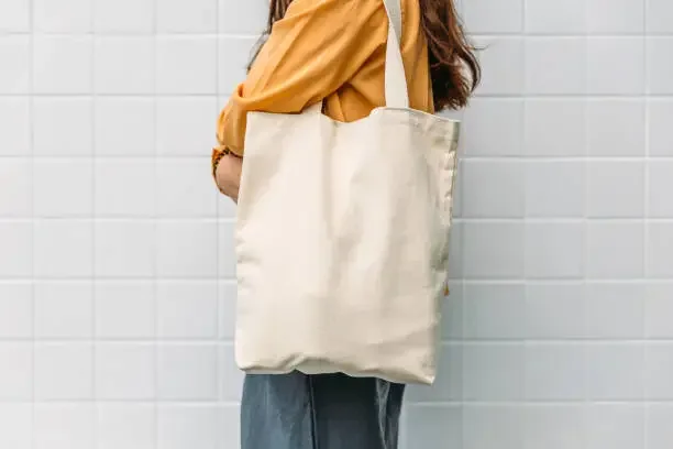 Person wearing a yellow jacket carrying a large white tote bag over their shoulder, standing against a white tiled wall. 