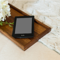 A Kindle e-reader on a wooden tray next to a white flower arrangement on a textured white brick wall background. Focus on Deception is also available on Kindle Unlimited.