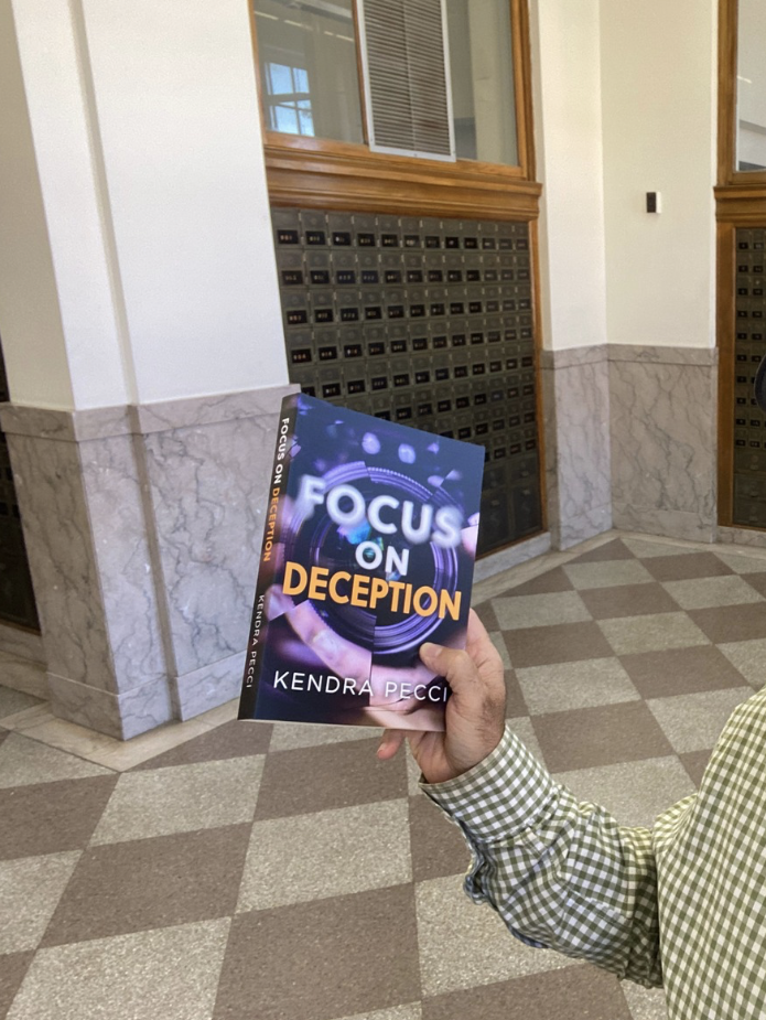 A Kindle e-reader on a wooden tray next to a white flower arrangement on a textured white brick wall background. Focus on Deception is also available on Kindle Unlimited.