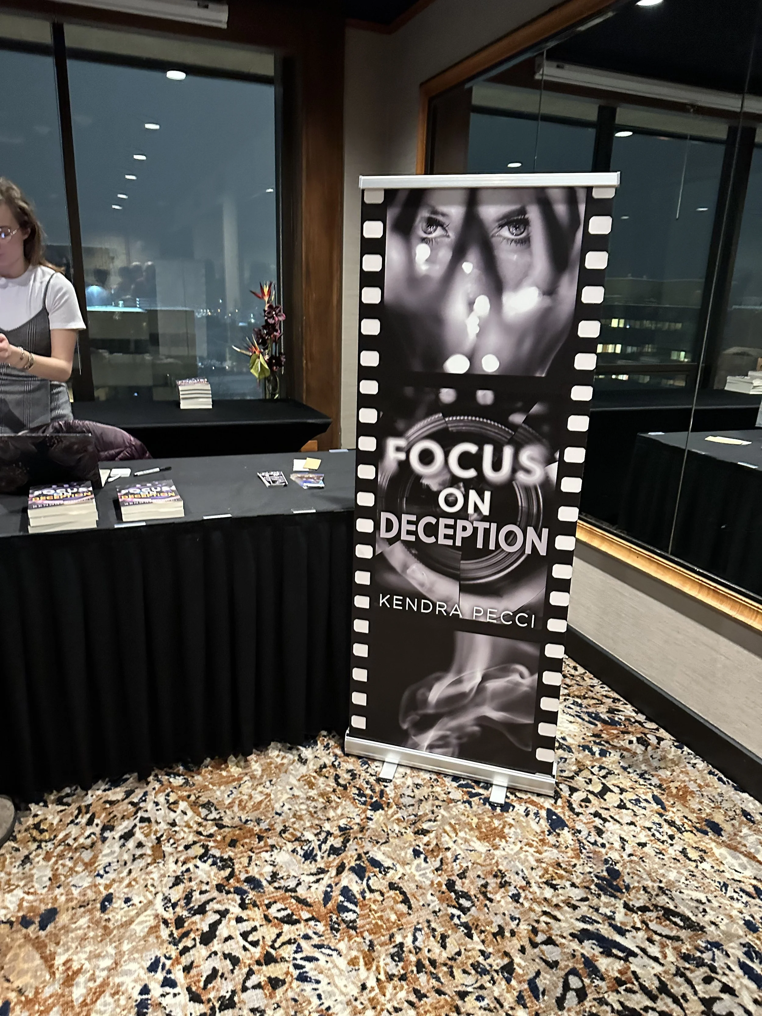 A young woman with dark hair sitting at a desk in a study area, reading a book under soft lighting with other people working nearby. Focus on Deception is a fast-paced read that is difficult to put down!