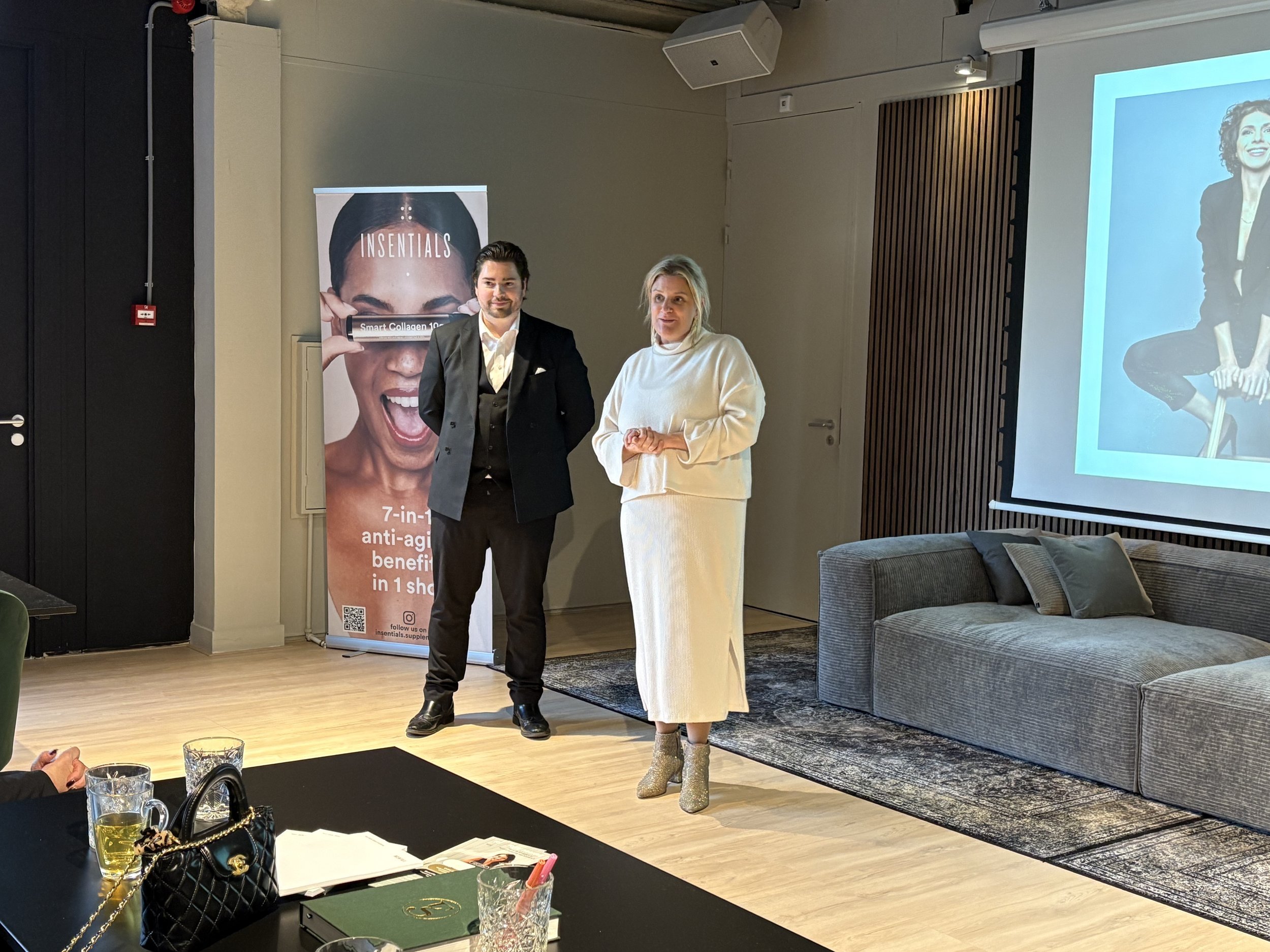 Two people standing in a room with a banner displaying skincare product information, next to a couch and a table with drinks and documents.