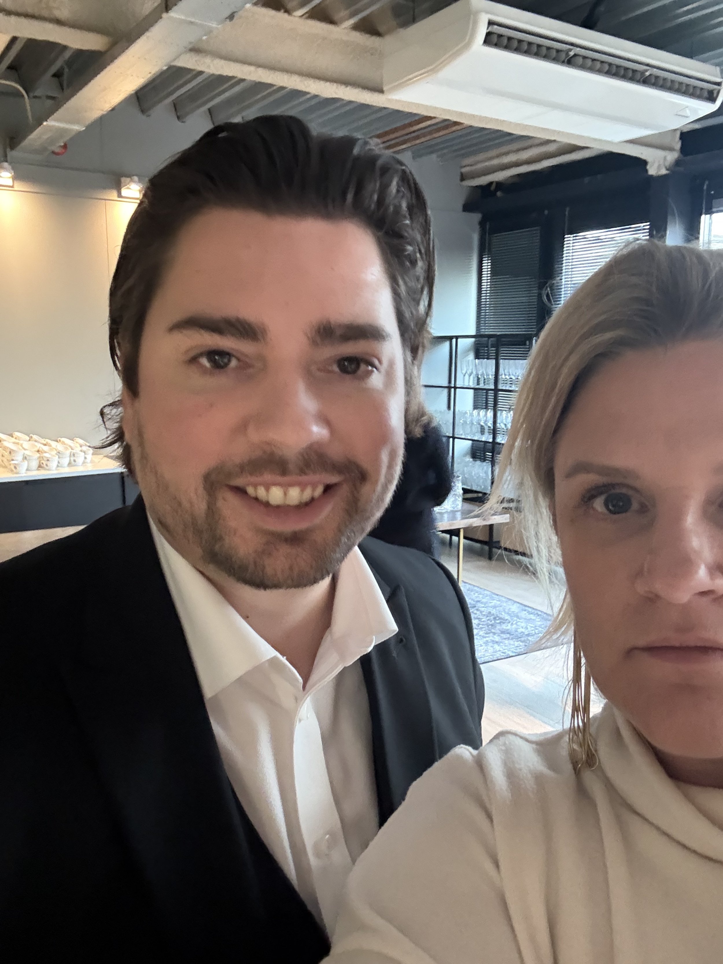 Two people posing for a selfie indoors with a modern ceiling and lighting in the background. The person on the left is wearing a black suit and white shirt, while the person on the right is wearing a light-colored top.