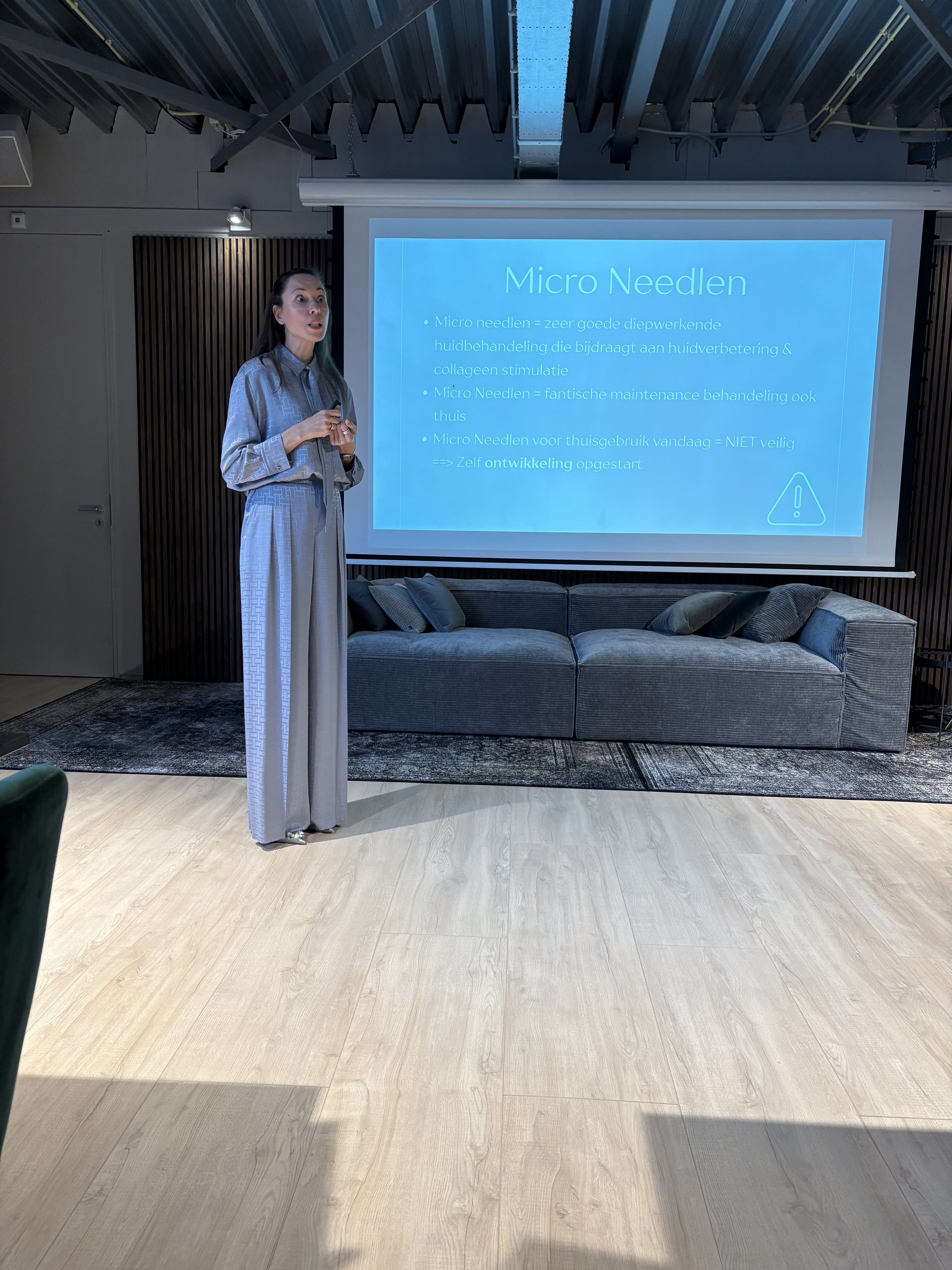 A person standing in front of a presentation screen inside a modern room. The slide on the screen is titled 'Micro Needlen' with bullet points in a foreign language. The room has a comfortable sofa, wooden flooring, and a high ceiling with exposed be