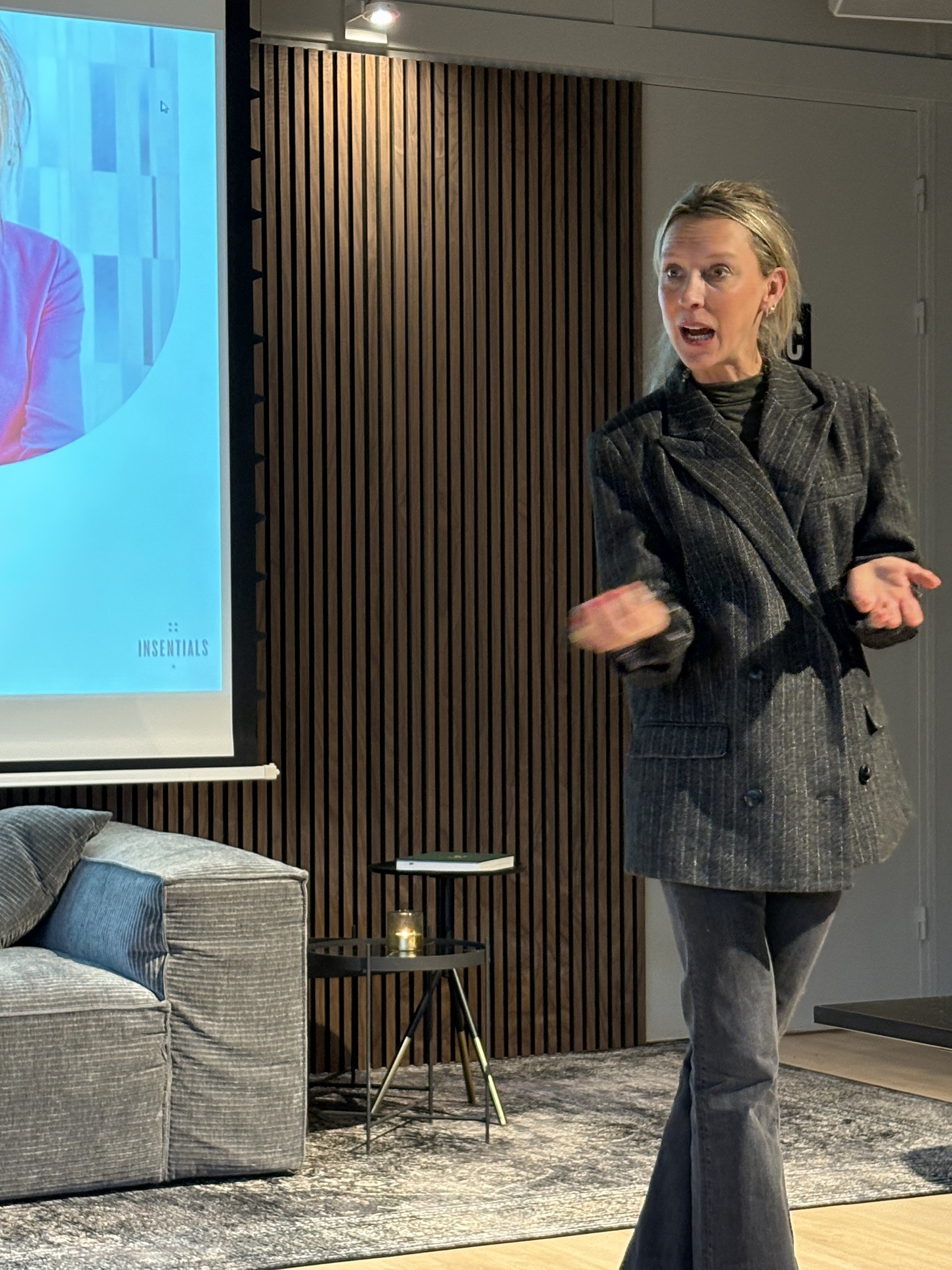 A person giving a presentation in a modern room with wooden paneling, standing near a gray sofa and a small table. They are gesturing with their hands and speaking. A screen displays a partial image and text. The setting appears professional and cont