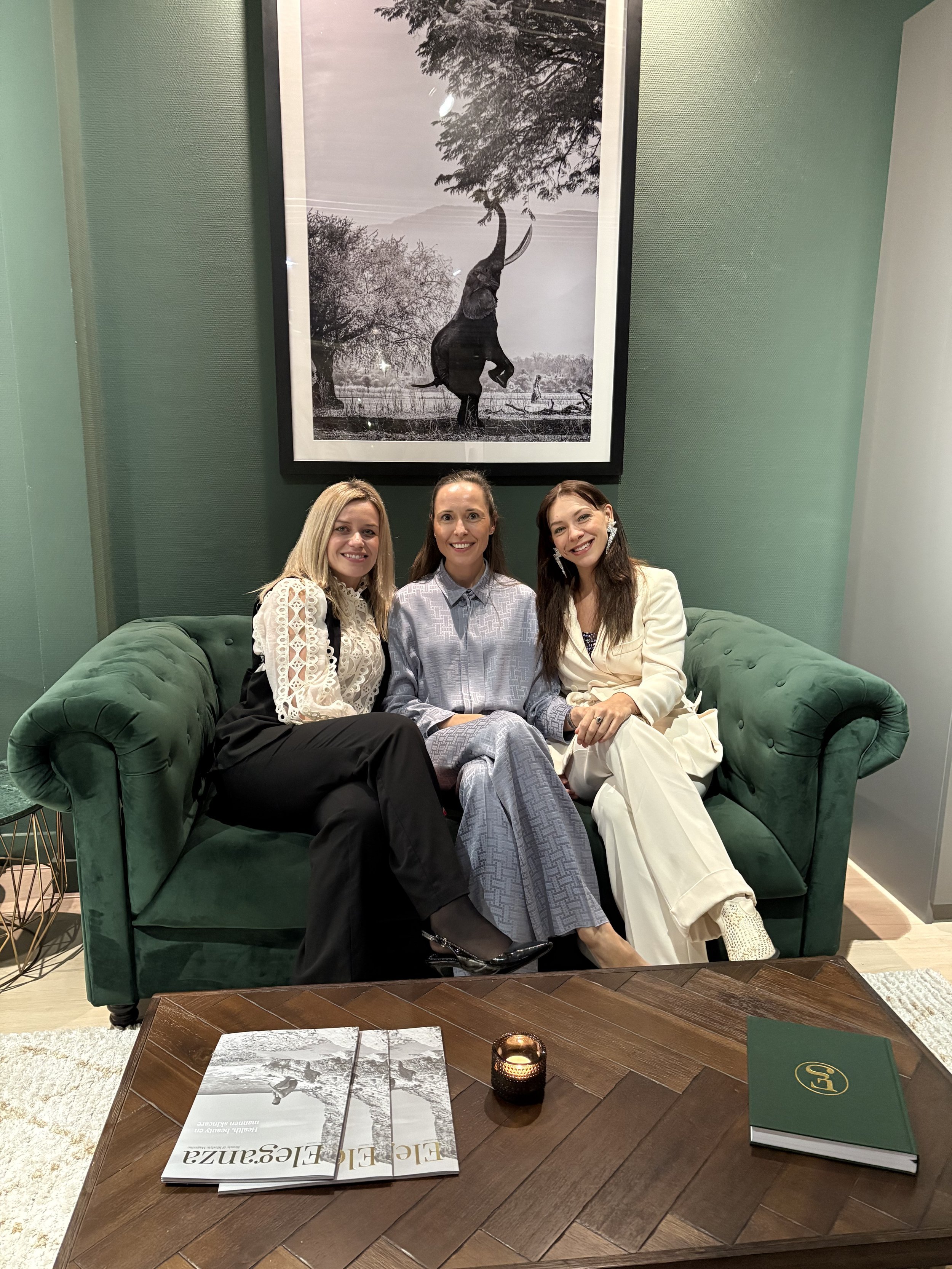 Three women sitting on a green sofa in front of a large framed photo of an elephant, with books and a candle on a wooden coffee table.
