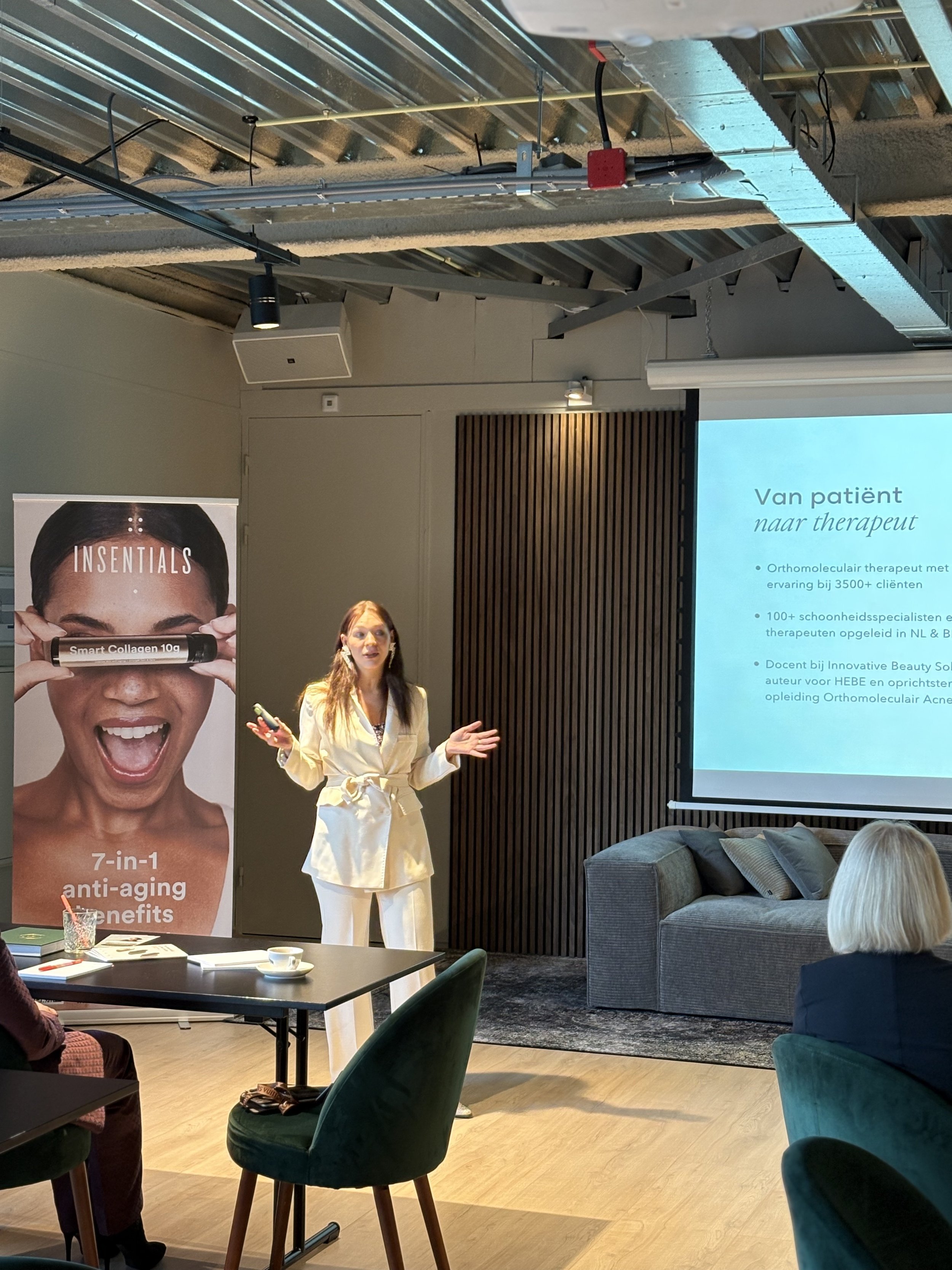 A woman in a cream suit is giving a presentation in a modern room with wooden floors. She stands near a projector screen displaying text in a foreign language. To her left is a banner advertising a product with '7-in-1 anti-aging benefits.' The room