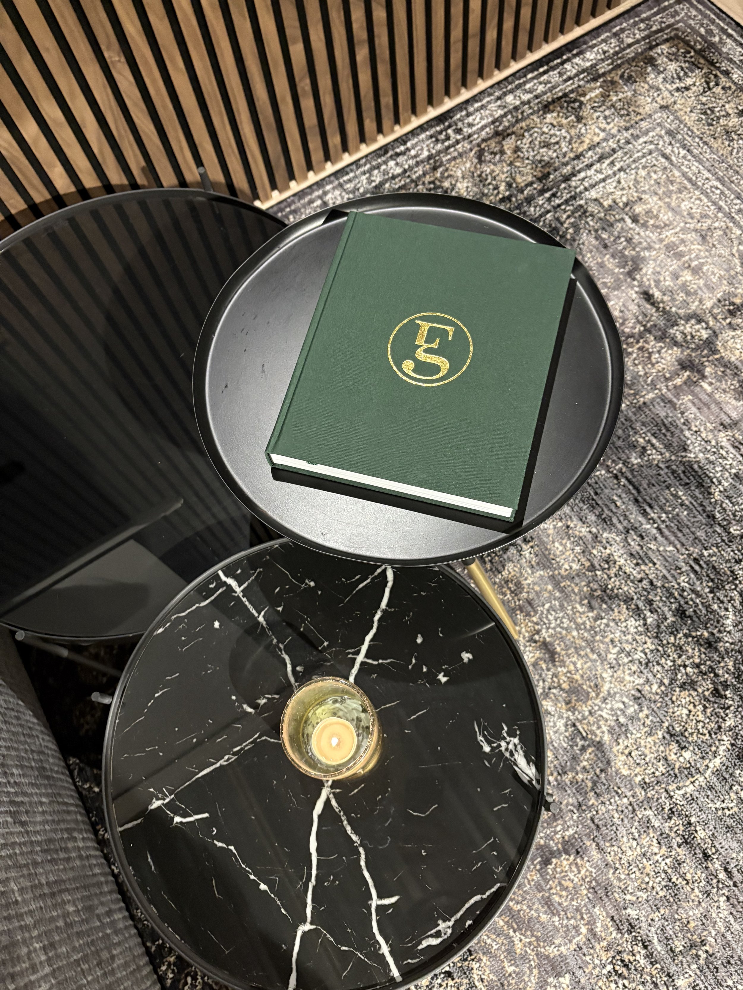 Green book with gold emblem on a round black table, next to a marble-top table with a candle, set on a patterned rug.