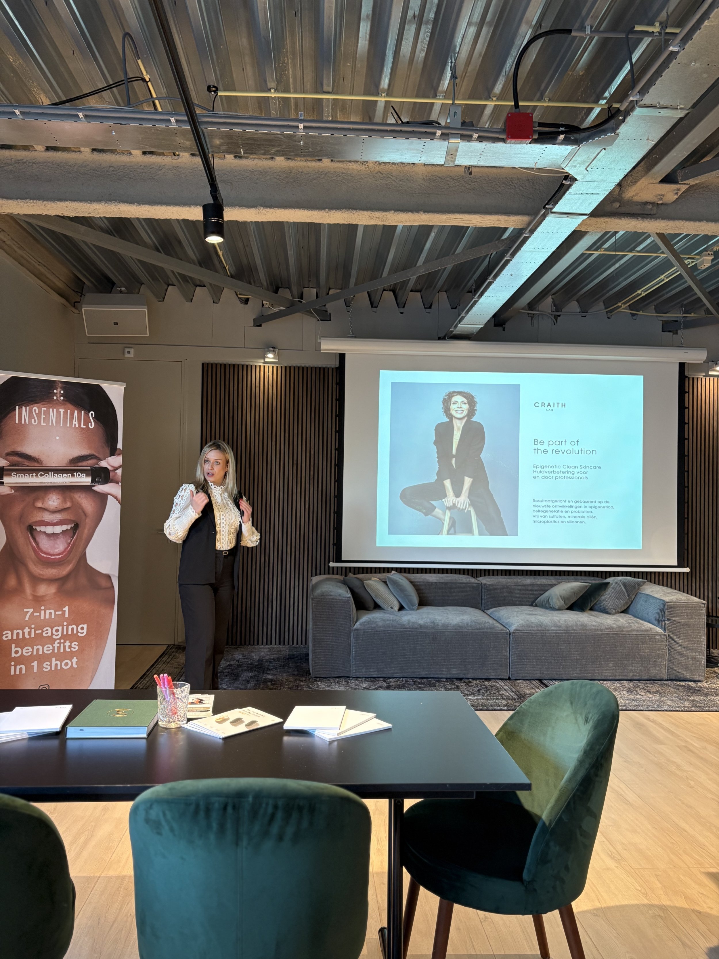 A woman presenting in a modern conference room with a screen displaying a presentation. There's a banner for "Insentials" featuring anti-aging benefits on the left. The room includes a black table with notebooks and a gray sofa in the background.
